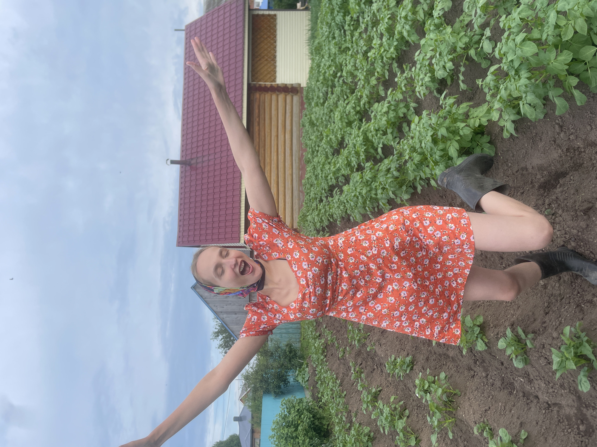 The media file shows a young woman in a red floral dress and black rubber boots, joyfully posing in a field of young plants, likely potatoes, in Kuyeda, Russia. Her arms are outstretched, and her mouth is open in a wide smile or shout of exultation. She wears a colorful scarf tied around her head, adding a rustic charm to her appearance. Behind her, a wooden house with a red roof and a section of light-colored siding is visible. The setting appears to be a rural homestead, with rows of cultivated plants stretching into the background under an overcast sky. The scene suggests a moment of freedom and happiness, perhaps during a harvest or a break from farm work. The time of day is indeterminate due to the cloud cover, but the overall atmosphere is cheerful and lively. There is no visible text in the media file.