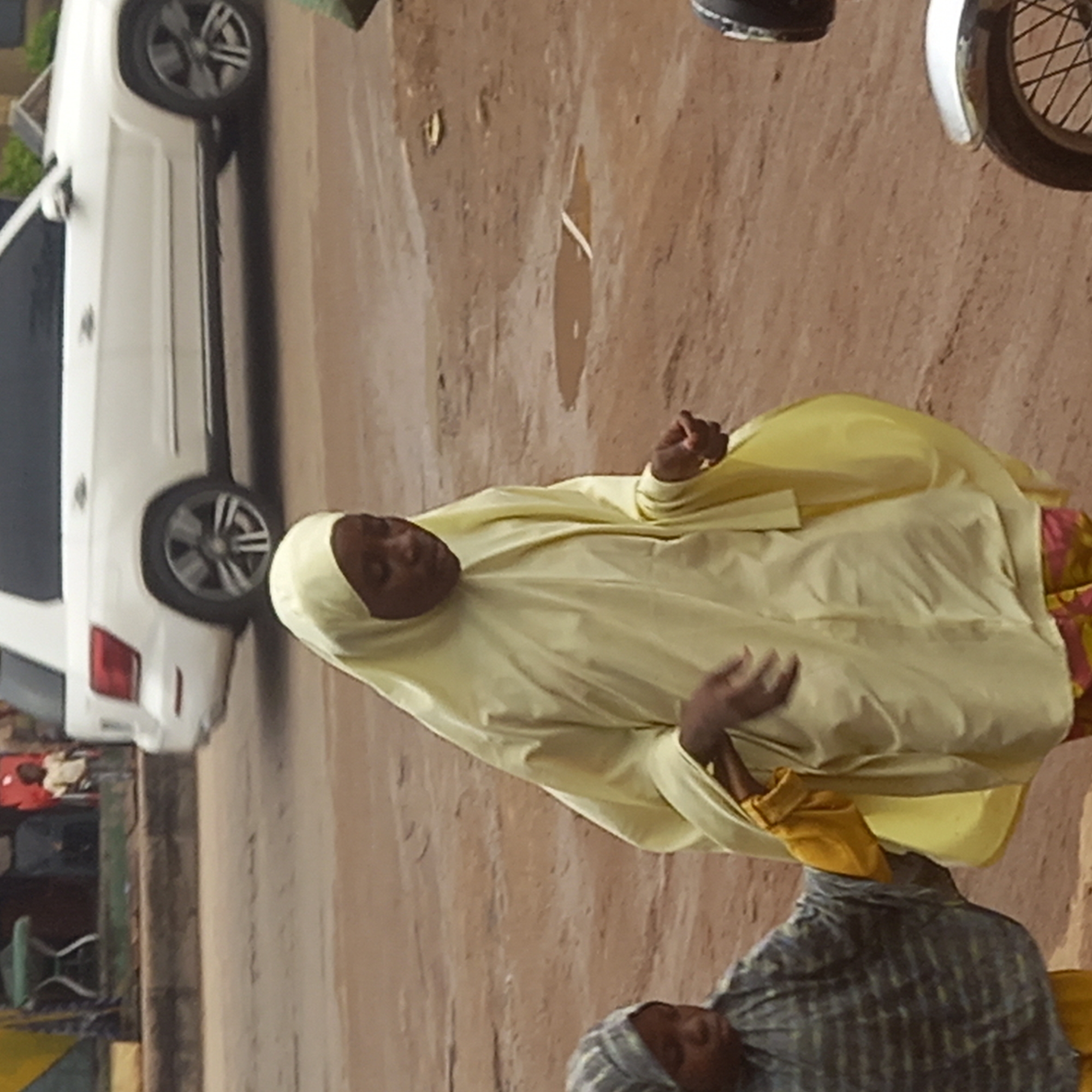 A woman wearing a light yellow hijab stands on a dirt or paved street in the city of Dadi Riba, Nigeria. A white car is parked to the left of her, and a motorcycle is parked to the right of her. Another person wearing a gray head covering is partially visible in the bottom center of the frame.