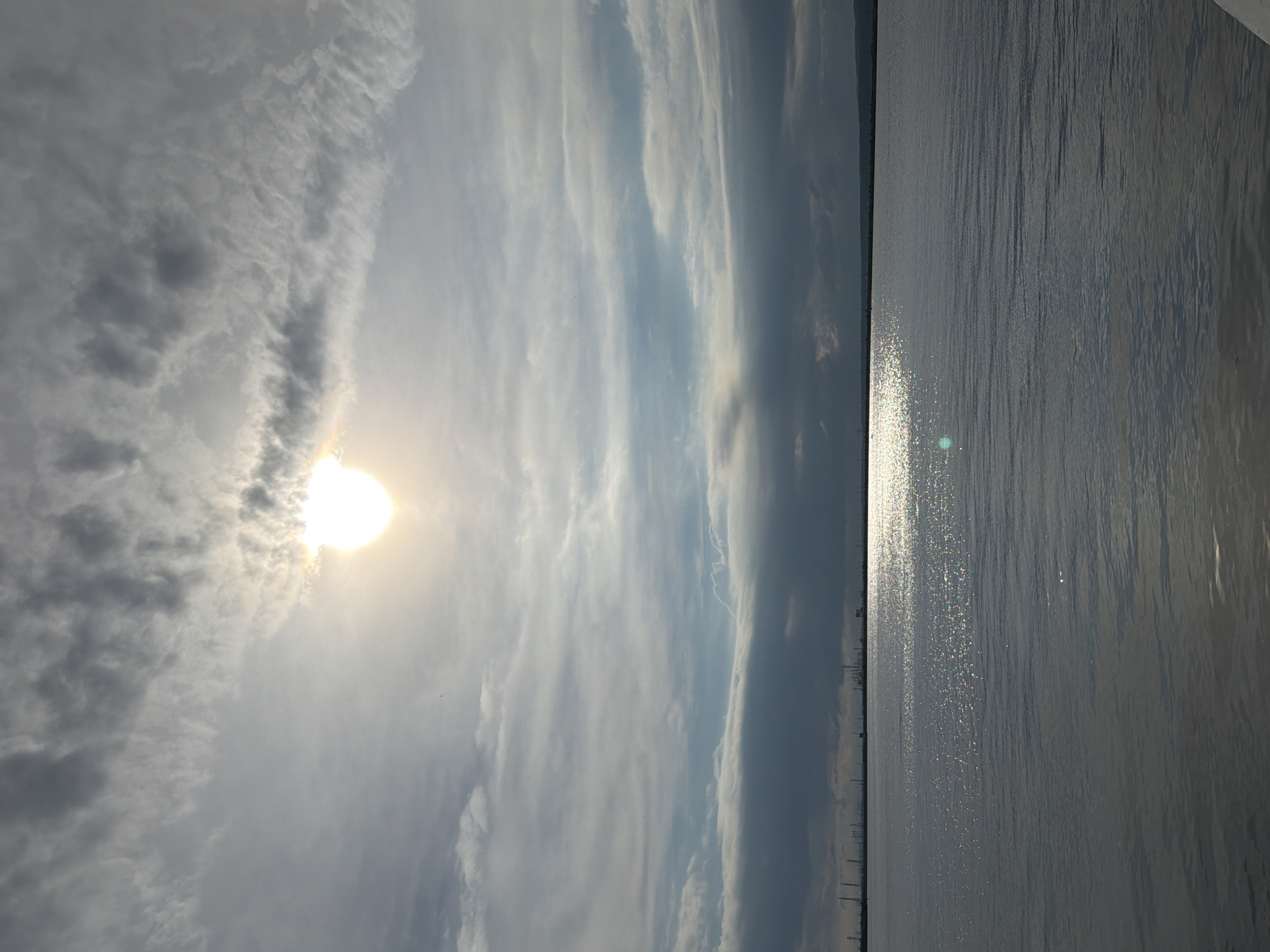 This is a photograph of the Kama River in Chaikovsky, Russia, taken from a low angle, likely from a boat or the shoreline. The sun is partially obscured by clouds, casting a bright glare on the water's surface. The sky is a mix of light and dark clouds. In the distance, along the horizon, are industrial structures, possibly a power plant, with tall smokestacks visible. The water is calm with gentle ripples, reflecting the light and the overcast sky. The overall mood of the image is serene and slightly dramatic due to the interplay of light and shadow.