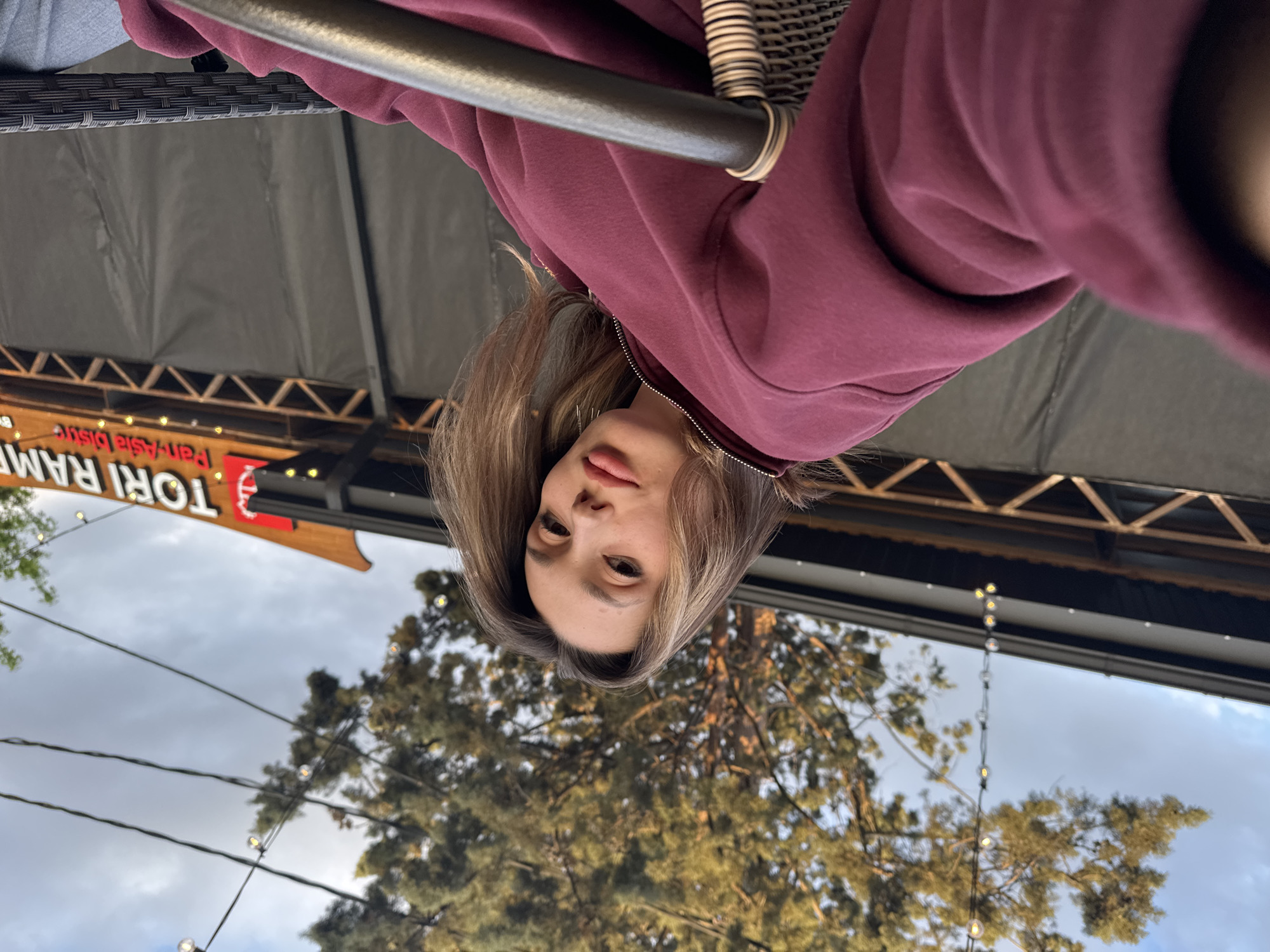 The image, when correctly oriented, displays a young woman looking up and posing for a photograph in an outdoor restaurant setting. She has long, light brown hair with blonde highlights and wears a maroon or burgundy full-zip hooded sweatshirt, partially zipped. Her expression is neutral and calm.
In the foreground, a dark metallic bar or railing runs horizontally across the top of the frame, behind which a dark woven chair back is visible. Above the woman, a dark canvas canopy or umbrella provides overhead cover.
The background features a cloudy or overcast sky, with the tops of several trees with green and brown foliage visible. String lights with small bulbs are strung across the scene, contributing to an outdoor ambiance. A wooden sign is prominently displayed in the background, featuring the text "TORI RAMEN" in large letters, and below it, "Pan-Asia Dishes", indicating the type of restaurant. The scene suggests an outdoor dining area, possibly a patio, on an overcast day or early evening.