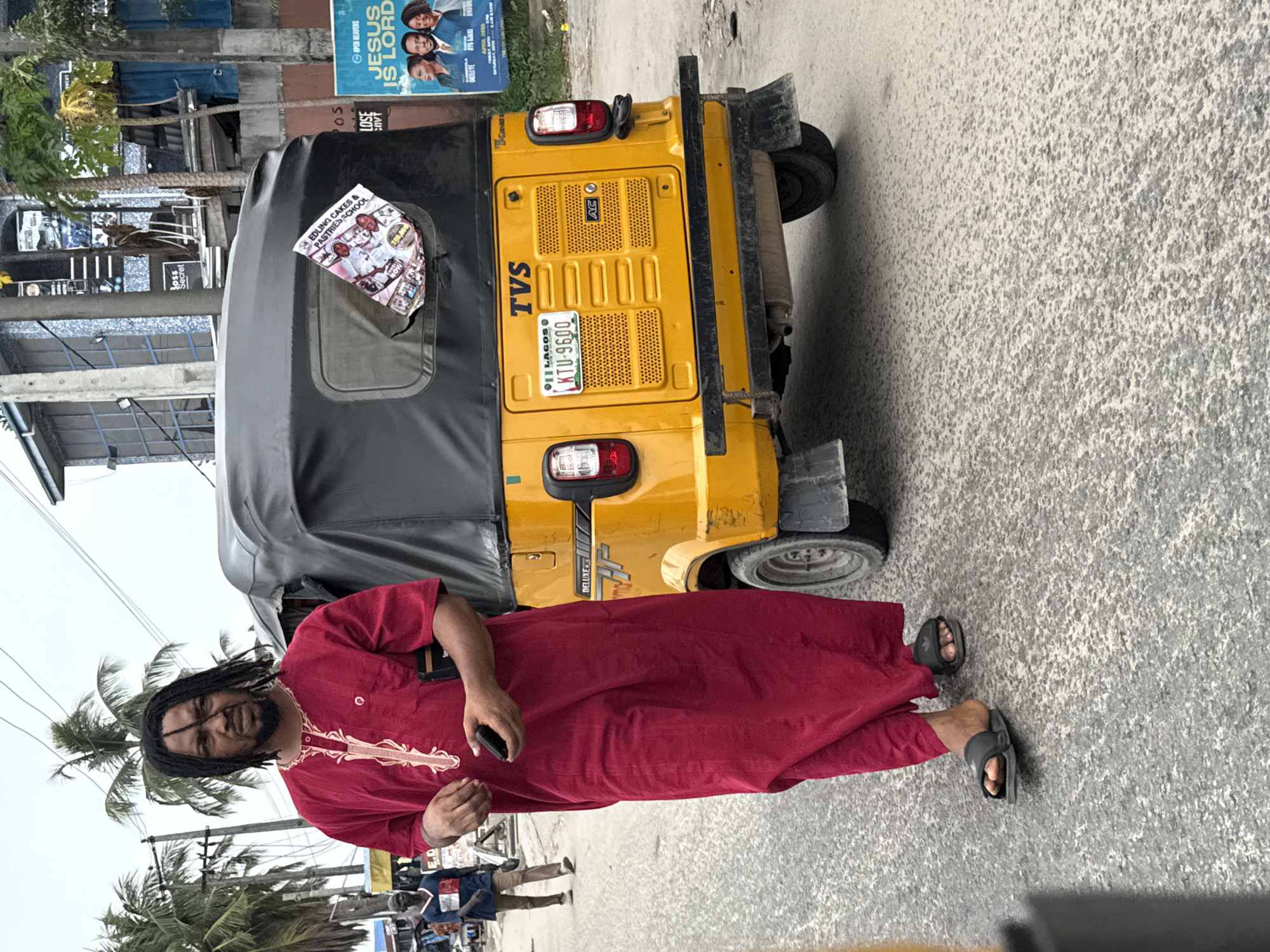 A man with dreadlocks, wearing a red robe and sandals, stands next to a yellow TVS tricycle with a black canvas roof. The tricycle's license plate reads "KTU-9600" and indicates it's registered in Lagos. A torn flyer advertising "EDLING CAKES & PASTRIES SCHOOL" is attached to the tricycle's rear window. In the background, a faded billboard promoting "JESUS IS LORD" is visible. The scene appears to be a street in a tropical location with palm trees and other pedestrians present. Based on the signage and context, the location is likely Ado, Nigeria.