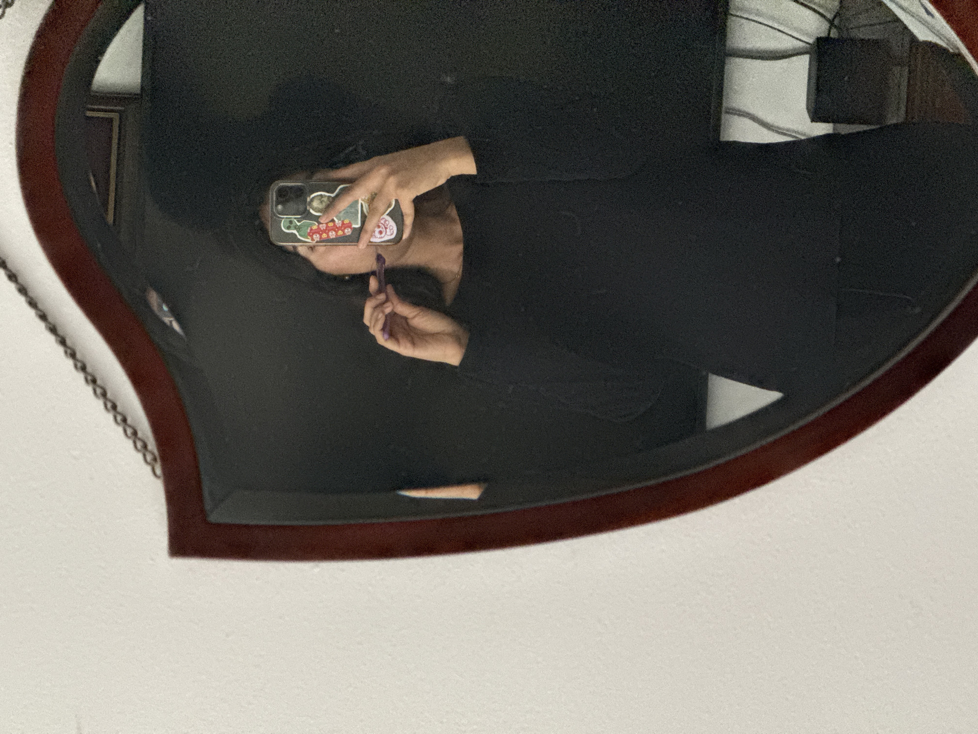 A person wearing a black long-sleeved shirt is reflected in a heart-shaped mirror with a dark brown wooden frame. The person is holding a smartphone with stickers on the back to take a selfie. They are applying lipstick. The mirror hangs on a white wall. Behind the person in the reflection, there's a glimpse of electronic devices, including wires and a black square box. The location is not discernible from the provided image to determine if it is in Puebla, Mexico.