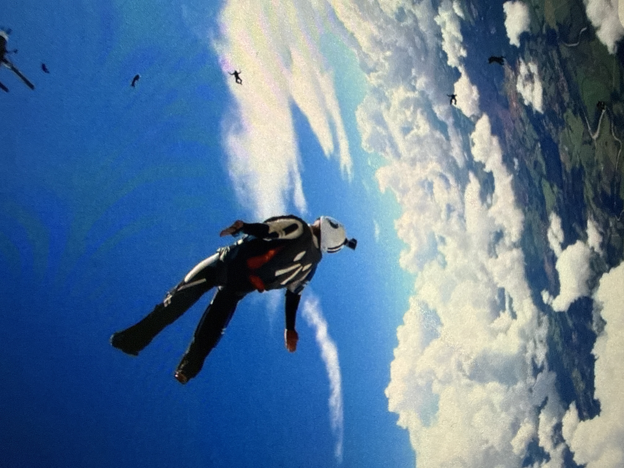 The image depicts a group of skydivers in mid-air. A primary skydiver is prominently positioned in the foreground, wearing a white helmet and a dark jumpsuit with white accents. Several other skydivers are scattered in the background at varying distances. Below them is a landscape interspersed with clouds. The location and city are undetermined.