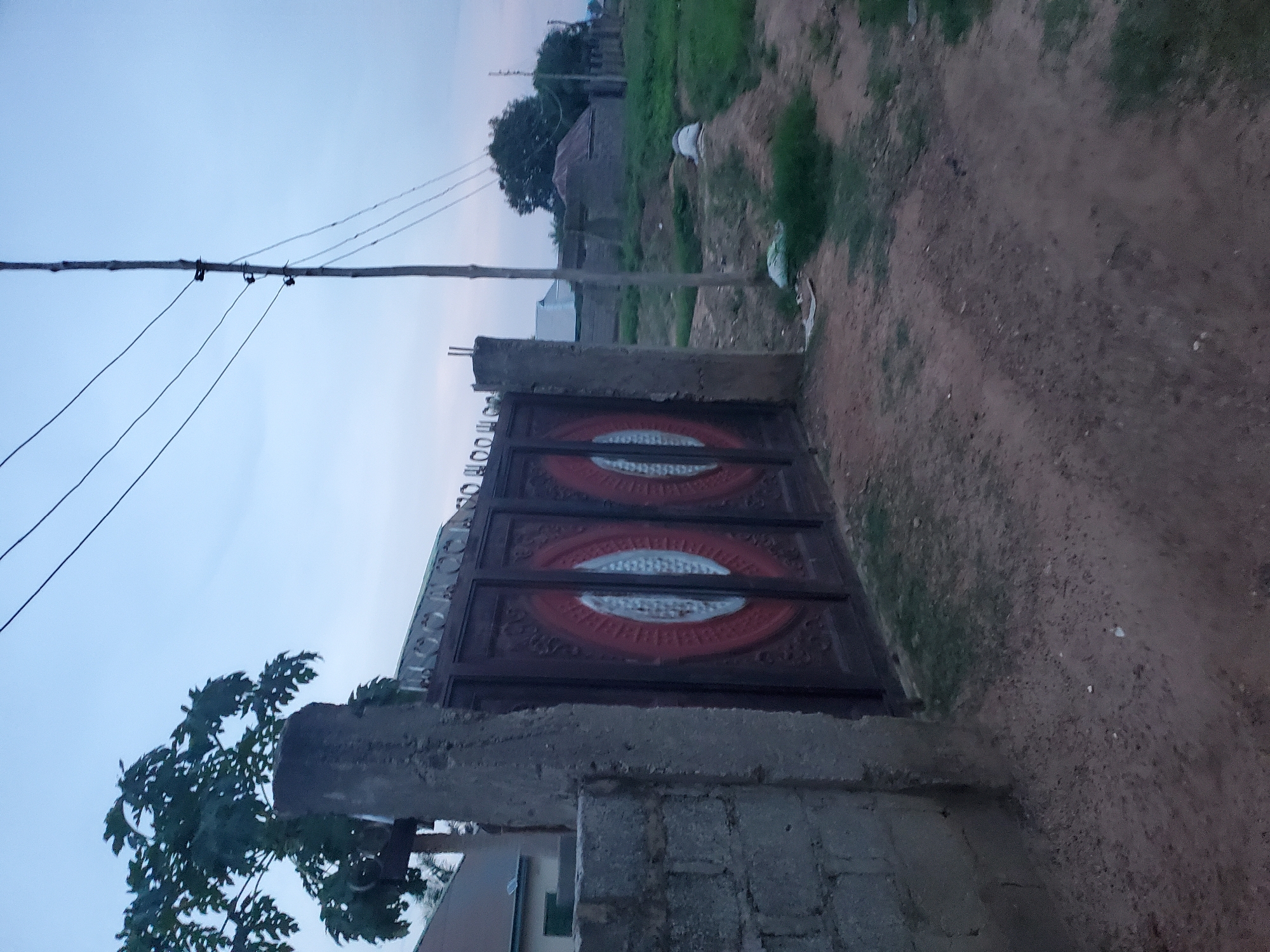 The image shows a view of a metal gate with decorative circular patterns, set between two concrete pillars. To the left of the gate is a brick wall. Behind the gate, there are various structures, including houses with sloped roofs and some smaller, possibly outbuilding-like structures. A utility pole with power lines runs vertically through the scene, extending into the sky. The ground in the foreground and middle ground is a mixture of dirt and sparse grass. The sky is overcast and appears to be dusk. There are no people visible in the image, and no actions or interactions are depicted. The location is described as the city of Winters, United States of America.