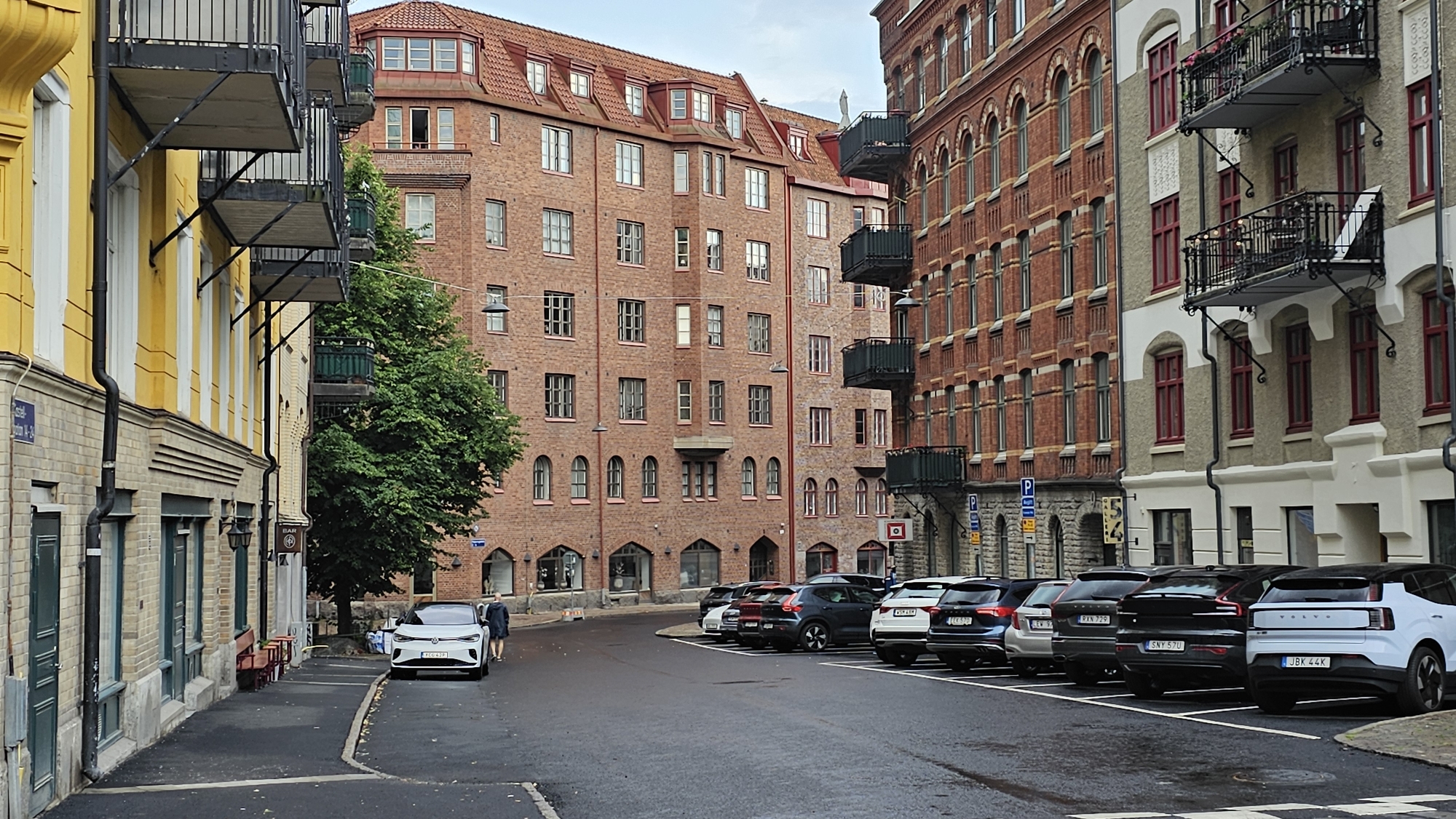 In Gothenburg, Sweden, on a cloudy day, a street scene unfolds with characteristic brick buildings. On the left, a yellow building with white trim features balconies with black railings and dark doors. To its right, a large, red brick building rises, adorned with numerous windows and balconies. Across the street, another red brick building with similar architectural elements stands, contrasting with a lighter-colored building featuring red-trimmed windows and balconies.
The street itself is wet, suggesting recent rain, and is lined with parked cars, including a white Volvo, a dark SUV, and other vehicles. A person in a dark jacket and pants walks away from the white Volvo towards the parked cars. Street signs are visible, including parking indicators. The presence of trees, particularly a large green tree on the left, adds a touch of nature to the urban landscape. The overall impression is one of a well-maintained urban environment with a mix of historical and modern architecture.