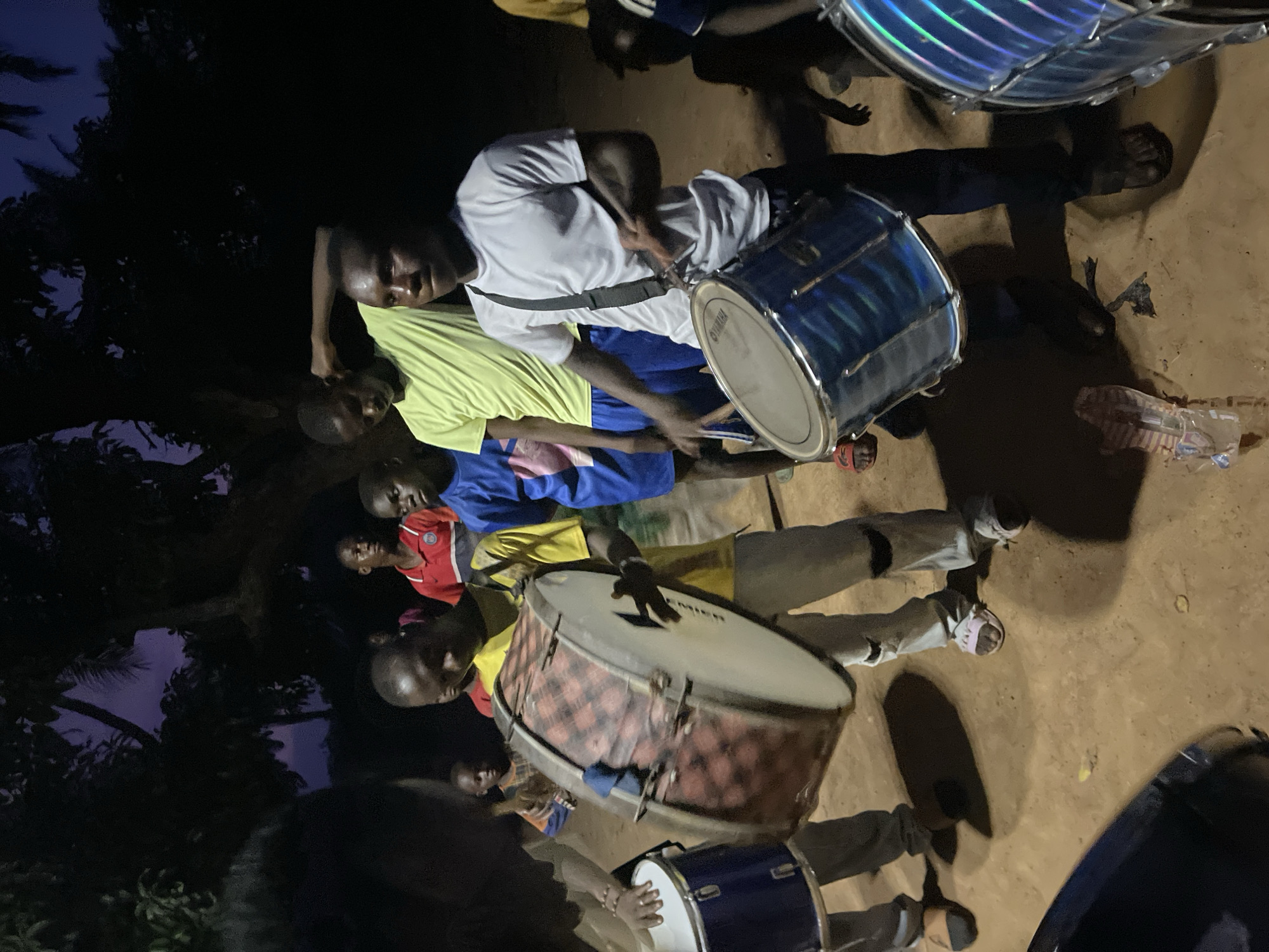 This image, taken at night in Andaha, Nigeria, depicts a group of young males and children gathered outdoors for a drumming session. The scene is illuminated by an unseen light source, casting distinct shadows on the dirt ground.
Several individuals are actively playing drums. A prominent figure in a white t-shirt is engaged with a large blue drum, which has "YAMAHA" visible on its head. Nearby, another person in a yellow t-shirt plays a large drum adorned with a red and black checkered pattern on its body and the brand "PREMIER" on its head. Other drums are also present, including a large one with reflective blue/purple stripes at the top and a smaller blue drum in the lower right. Most participants appear to be holding drumsticks.
The dark sky and shadowy environment confirm that the event is taking place at night or late dusk. Dark trees and foliage are visible in the background. The participants are casually dressed in t-shirts and trousers or shorts, with some wearing sandals. The gathering conveys a sense of communal musical activity or practice. A plastic container can be seen on the ground in the upper right portion of the image.