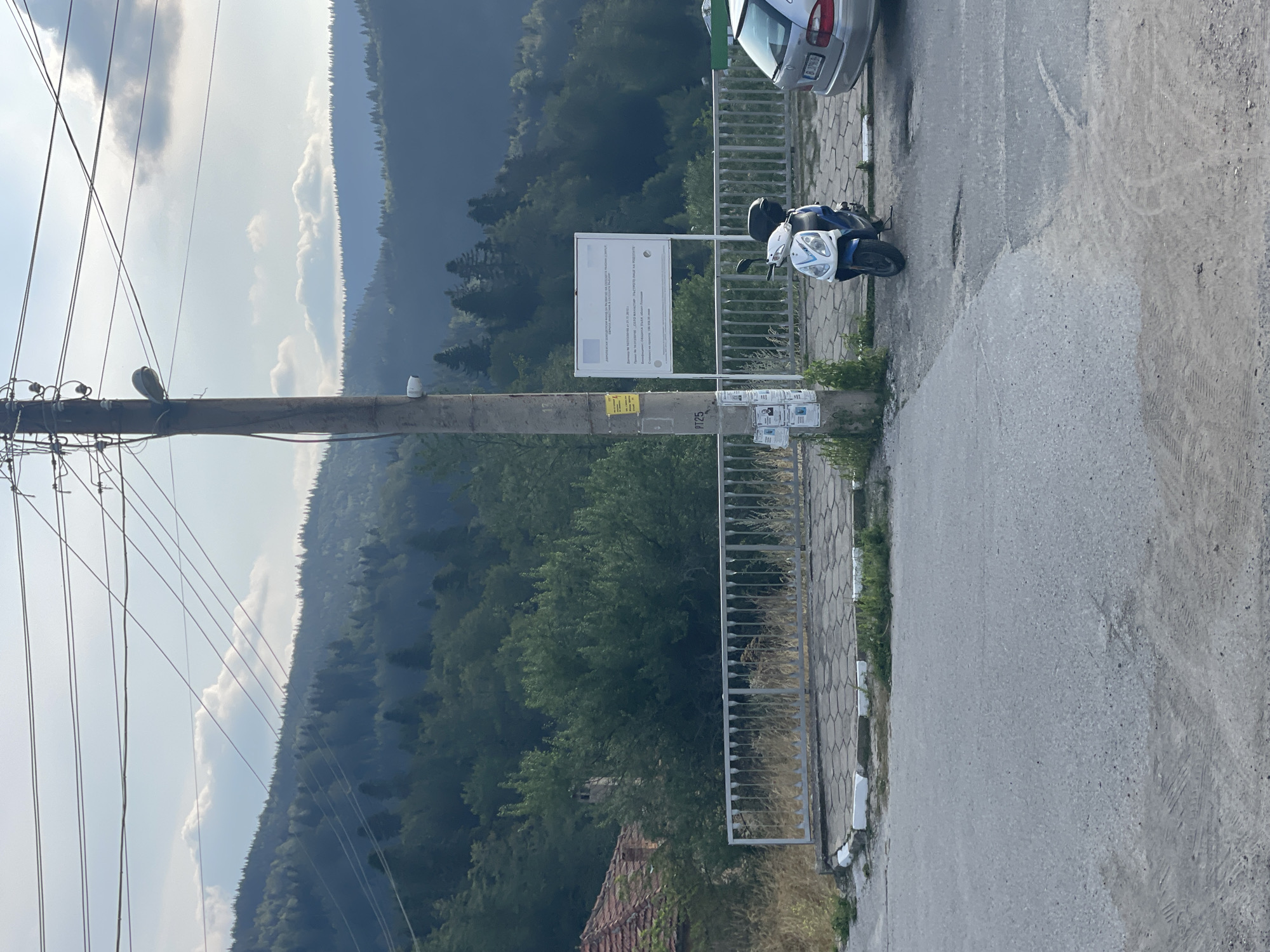 The image depicts a rural or semi-rural roadside scene with a mountainous, forested backdrop. A utility pole stands prominently in the foreground, adorned with wires, a lamp, a security camera, and various posters. To the right of the pole, a white and blue scooter is parked on a paved area, next to a silver sedan. A metal fence runs along the paved area, separating it from an area with overgrown grass and what appears to be a tiled roof of a building visible on the left. The background consists of densely wooded hills under a partly cloudy sky. The overall impression is one of a quiet, perhaps slightly weathered, location. The context provided suggests this scene is in Manastir, Bulgaria.