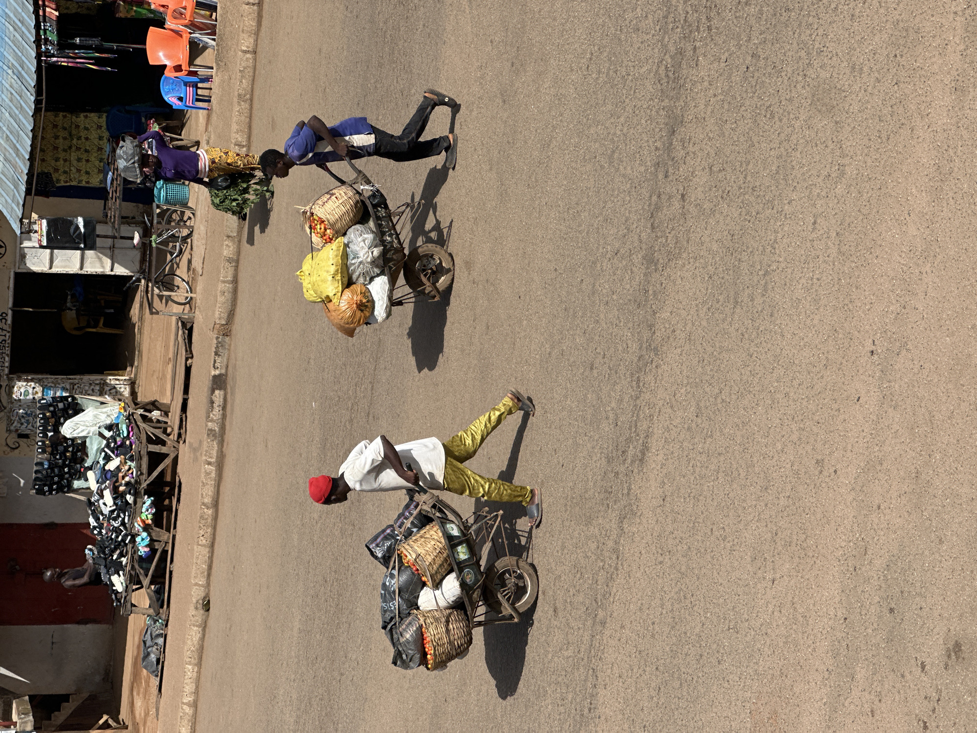 In a street scene in Bukuru, Nigeria, two individuals are pushing loaded wheelbarrows. The first person, wearing a red cap and a white t-shirt with yellow pants, pushes a wheelbarrow filled with baskets of produce, bags, and other goods. To the right, a second individual, in a blue shirt and black pants, pushes a similar wheelbarrow containing baskets of produce and sacks. In the background, a market stall displays a variety of sandals, and several people are visible near buildings and more market stalls. Chairs are set out in front of one of the establishments. The ground is paved asphalt, and the overall scene depicts a bustling marketplace environment.