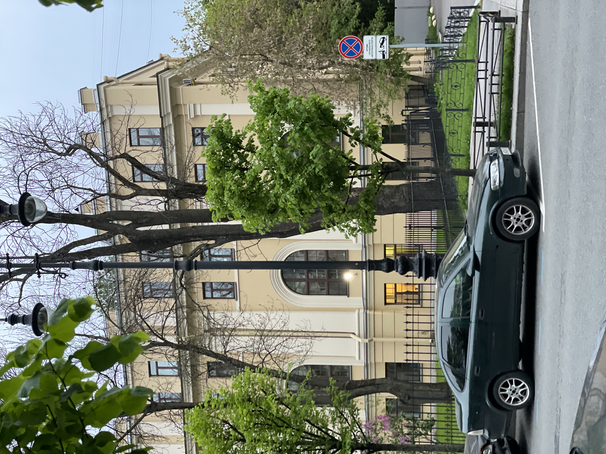 The image shows a street scene in Saint Petersburg, Russia. A dark green car is parked along the side of the road, partially blocking the view of a light yellow building behind it. The building is several stories tall and has numerous windows. A black wrought-iron fence runs along the edge of the building. Several trees are present, some with green leaves and others with bare branches. A street lamp is visible in the foreground. A street sign indicating no parking is also present. The sky is overcast.