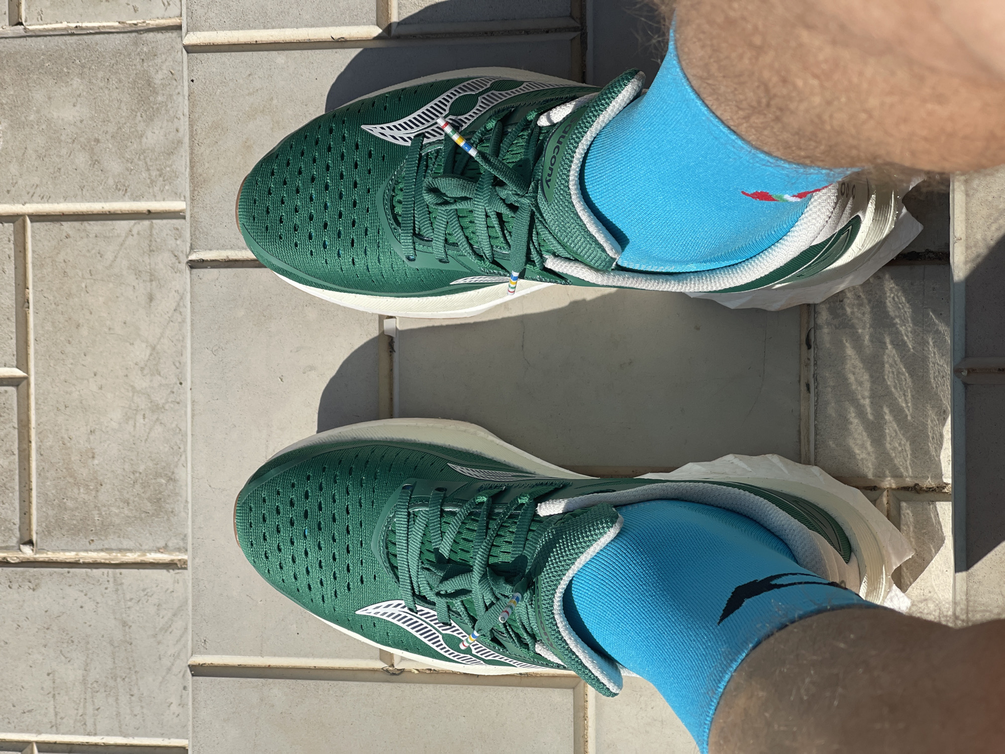 A top-down shot shows a person wearing a pair of green Saucony running shoes with white soles and white Saucony logos. The shoes are laced with green laces that have small stripes of color on the ends. The person is also wearing bright blue socks that have a black logo on them. The person is standing on a tiled surface, possibly a sidewalk or patio. Shadows are visible on the surface, suggesting it is a sunny day. The location of Перевозное, Russia is not discernible from the image.