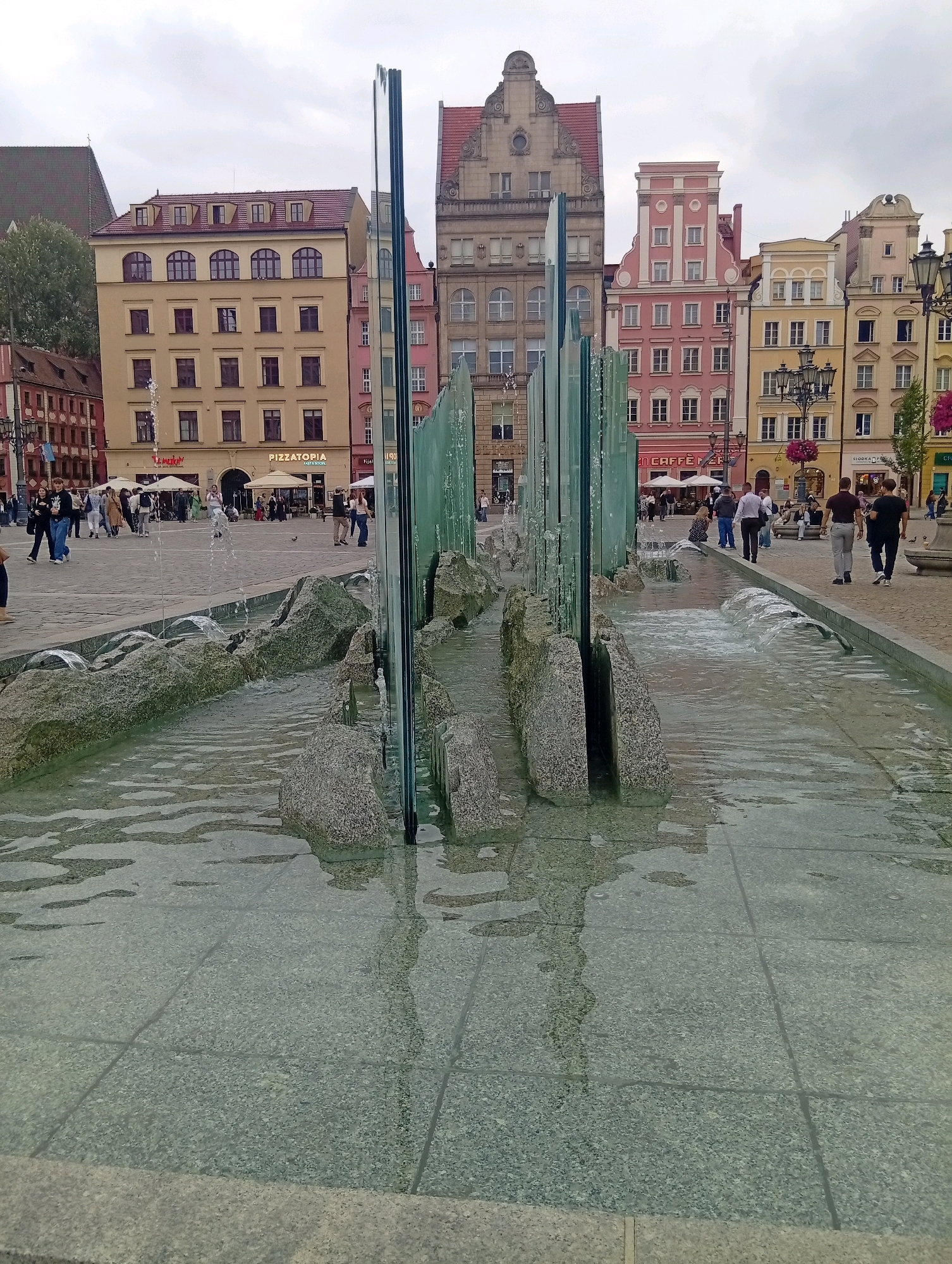 The image depicts a modern water feature in the Market Square of Wrocław, Poland. The foreground shows a shallow pool of water with a textured, tiled floor, bordered by rough-hewn rocks and tall, vertical glass panels. Water streams down the glass and flows around the rocks, creating a dynamic effect. In the background, a row of colorful, historic buildings with varied architectural styles lines the square. People are visible walking in the distance, some near outdoor seating areas of cafes and restaurants. The sky is overcast, contributing to a diffused light. The overall scene captures a blend of historical architecture and contemporary urban design in a bustling public space.