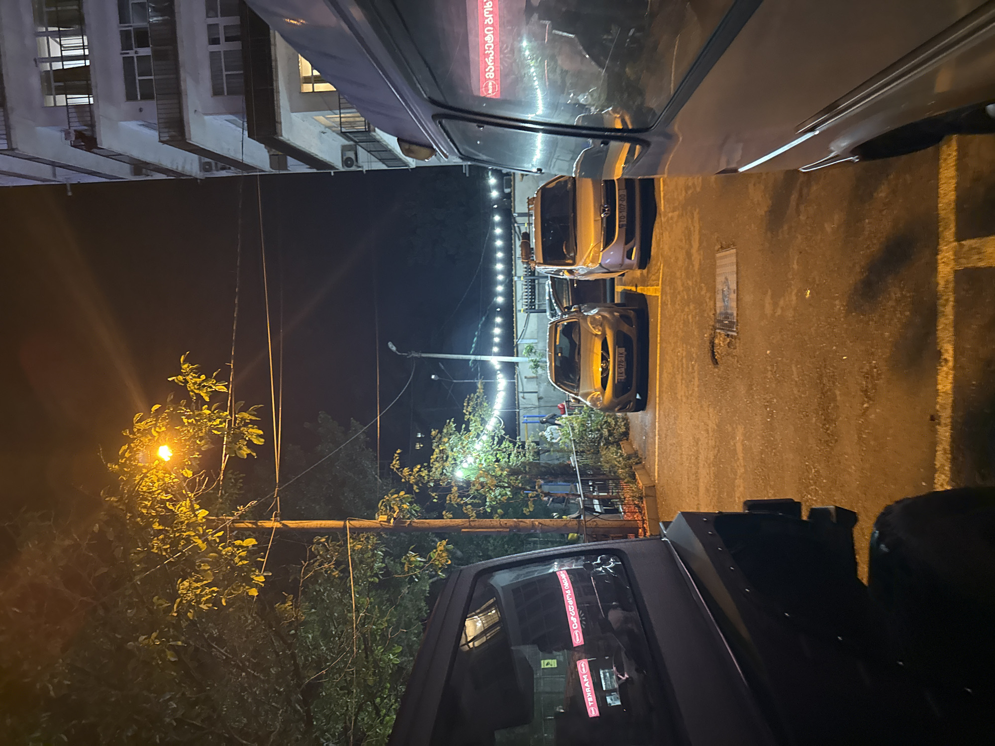 At night, a parking area is visible in front of a multi-story white apartment building, likely in Tbilisi, Georgia, given the Georgian script on the vehicles. The scene is illuminated by streetlights and string lights hung across a courtyard. In the foreground, the left side of a black SUV is parked, with red Georgian text visible on its rear window. To the right of the SUV, two cars, a silver hatchback and a light brown minivan, are parked in marked spots. A light purple van is parked on the far right, its side panel reflecting the ambient light. Beyond the cars, a metal fence separates the parking area from a landscaped section with trees and bushes. Several people are faintly visible in the background, near the fence and the building. The ground is asphalt, with yellow parking lines marking the spaces.