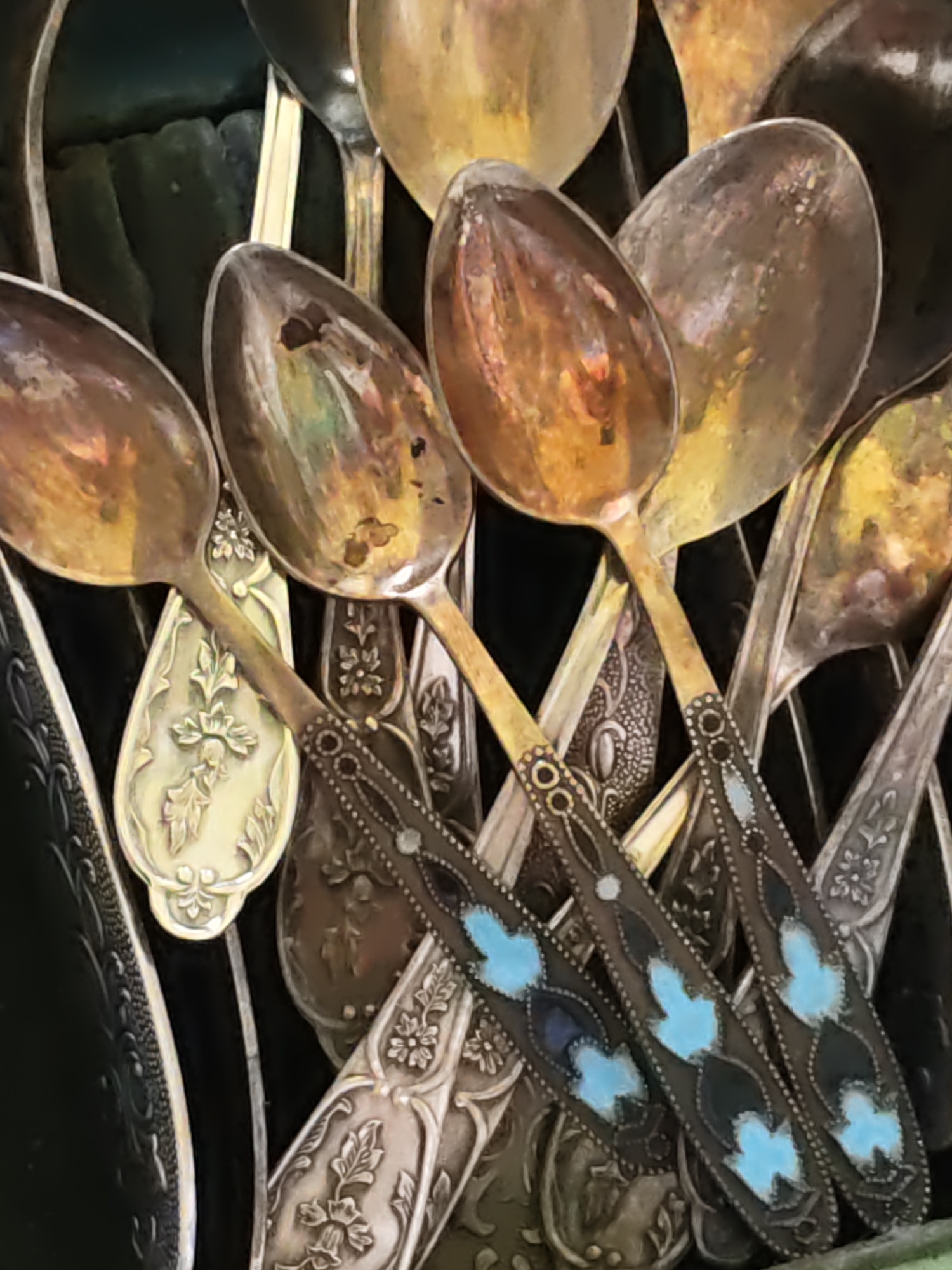 The image is a close-up, high-angle shot of a collection of antique spoons, likely an heirloom or decorative display. The spoons are made of various metals, with some appearing to be silver or silver-plated, showing signs of tarnish and age. They feature intricate engravings and decorative elements on their handles and bowls, including floral patterns and geometric designs. Some spoons have enamel inlays of blue and black. The spoons are arranged haphazardly, overlapping and layered on top of each other, creating a textured and visually rich composition. The lighting is warm and appears to be natural, highlighting the reflective surfaces and casting subtle shadows. The background is dark and out of focus, serving to emphasize the spoons themselves. The image does not depict any people, actions, or specific environmental context that would place it in Rostov-on-Don, Russia, or any other specific city.