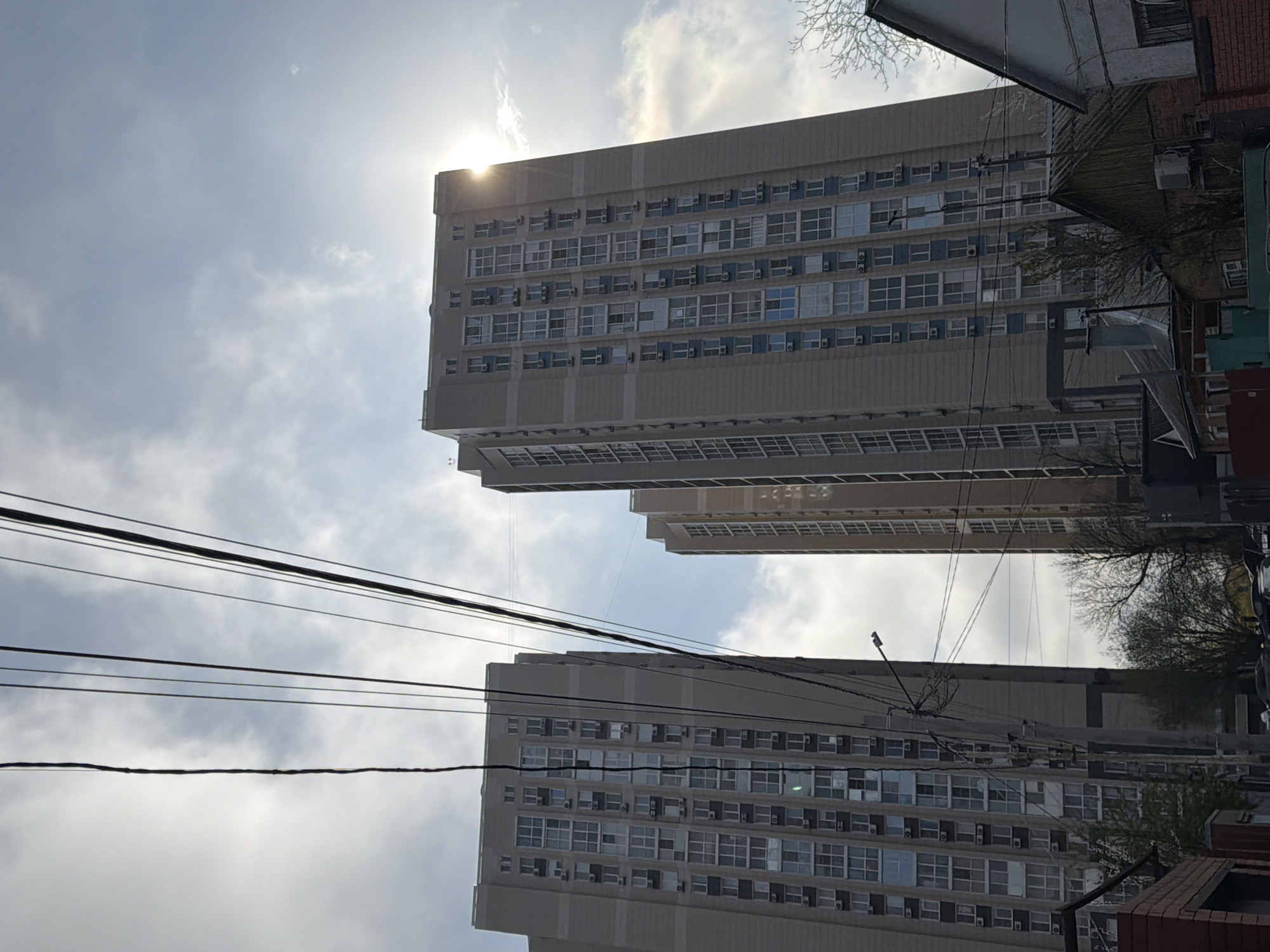 The image depicts two high-rise buildings in Rostov-on-Don, Russia, viewed from a low angle. The buildings are a beige color and have many windows with what appears to be air conditioning units in each. Power lines are visible in the foreground, and a portion of a tree is visible on the right-hand side of the image. The sky is cloudy, and the sun is obscured behind the top building. There are no people visible in the image.