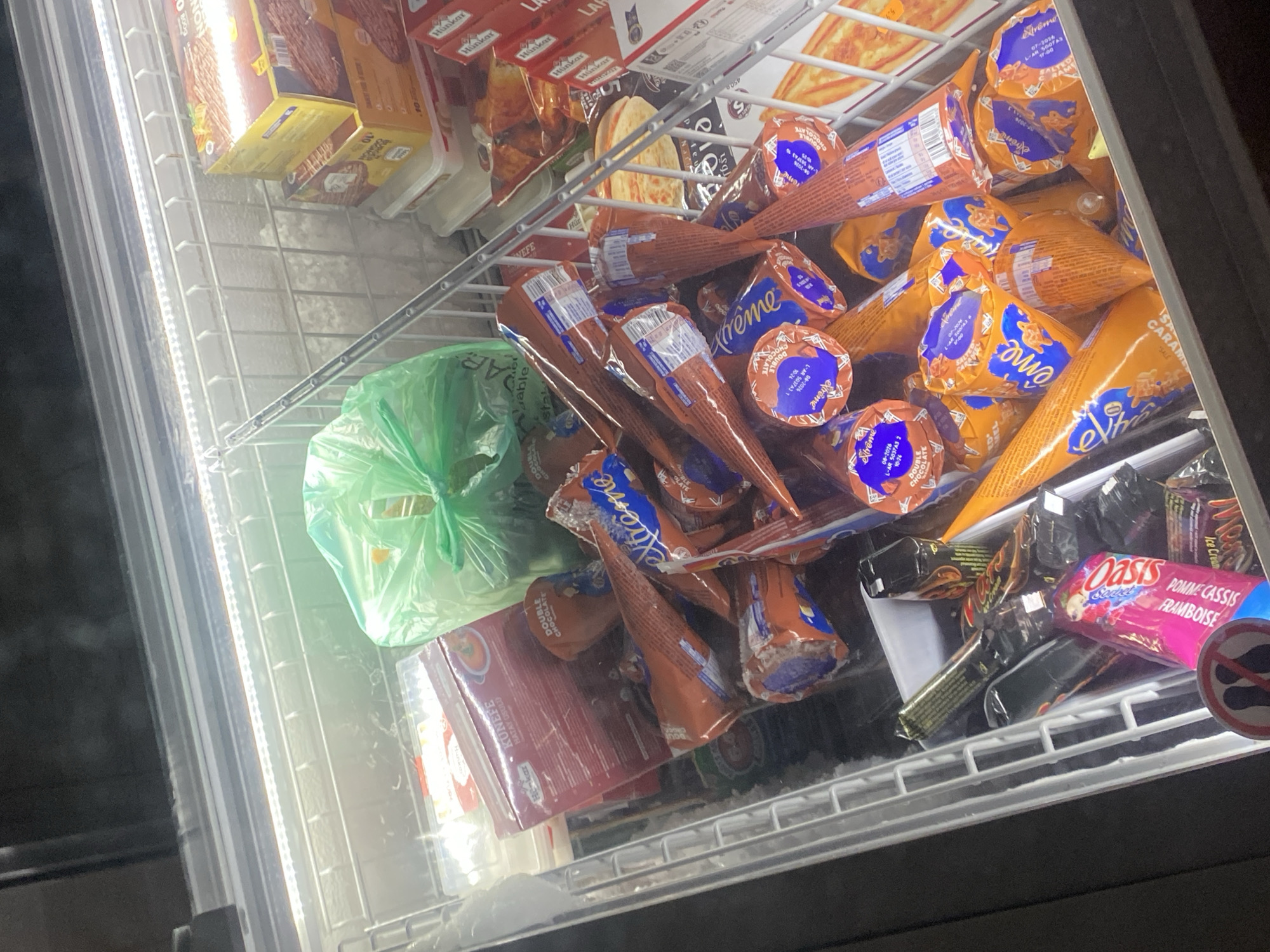 The image displays the illuminated interior of an open-top freezer display unit located within a retail establishment in Schiltigheim, France. The freezer contains a varied and somewhat disorganized assortment of frozen food products. Numerous individually wrapped ice cream cones, predominantly in orange packaging with blue accents, occupy the central and right sections. These cones are marked with the brand "Extrême" and include descriptions such as "Double Chocolate" and "Salted Caramel." In the upper left quadrant, a red box labeled "KUNEFE HATAY USULÜ" by "Hun-kar" is visible next to a crumpled green plastic bag. Further right, several stacked yellow and red boxes, some identified as "Hunkar" brand, contain other frozen goods, including visible pizza-style products. The bottom left area of the freezer holds a collection of individually wrapped confectionery or ice cream bars, specifically noting a pink-packaged "Oasis FRAMBOISE P. CASSIS" and a "Mars Ice Cream" bar. A partial circular sign, depicting a crossed-out footprint icon, is discernible at the extreme bottom left. No individuals are present in the scene.
