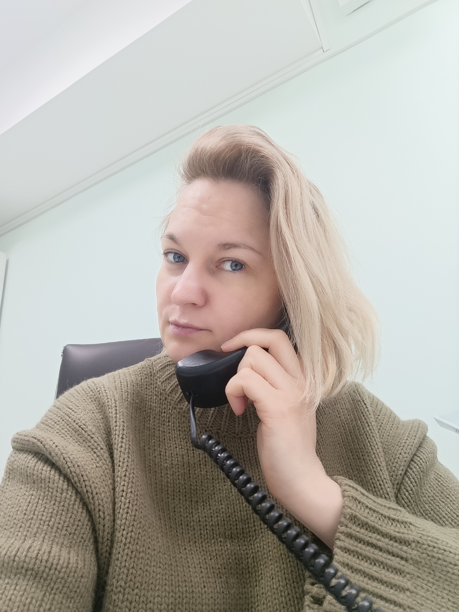 A woman with blonde hair and blue eyes is holding a black landline telephone receiver to her ear. She is wearing an olive green, knitted sweater. The background includes a light green wall and a white ceiling with a linear vent. A black office chair is partially visible behind her. The location is identified as Марковский, Russia.