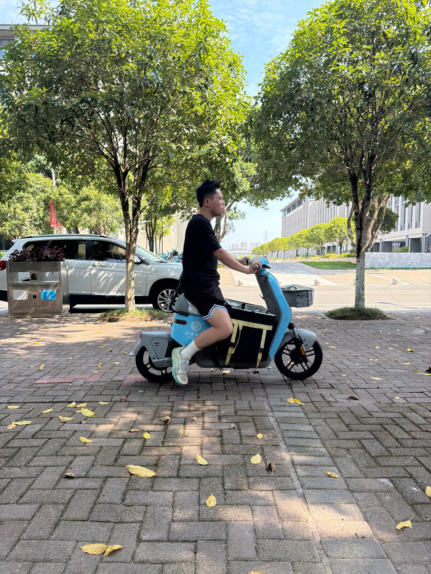 In Jiangjin, China, a young man rides a blue electric scooter along a tree-lined brick pathway. He is wearing a black t-shirt, black shorts with white stripes, white socks, and light-colored sneakers. The scooter has a grey seat and a black storage box attached to the front. Yellow leaves are scattered on the pathway. In the background, a white SUV is parked to the left, and modern buildings with large windows and manicured trees are visible. The sky is clear and blue.