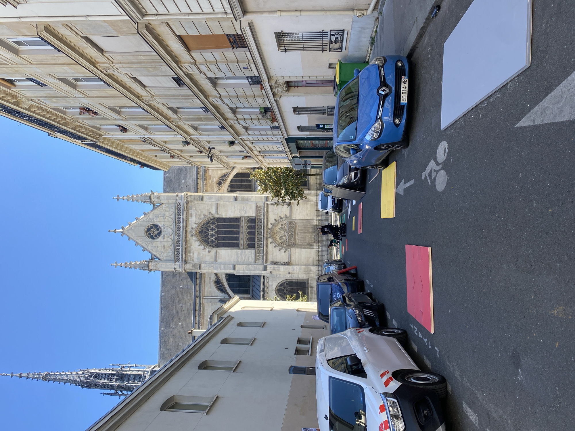 This media file captures a sunny street scene in Paris, France, looking towards a prominent Gothic church. The image is oriented vertically, showing the sky at the bottom and the street receding upwards.
On the left side of the street, a multi-story, light-colored building with ornate architectural details, numerous windows, and small balconies lines the path. On the right, another traditional building stands, with its ground floor featuring shopfronts and a tree growing in front of it.
The central focus is a grand Gothic church or cathedral, characterized by its intricate stone facade, rose windows, pointed arches, and decorative spires. Further in the background, a very tall, slender Gothic spire rises into the clear blue sky, suggesting another significant landmark nearby.
The street itself is asphalt, with several vehicles parked along the right side. A bright blue Renault Scenic car with the French license plate "EE-014-WB" is prominently visible. Behind it, several other dark-colored cars are parked. Closer to the foreground, a white delivery van is also parked.
Notable details on the street surface include rectangular panels of white, yellow, and red colors, possibly indicating temporary markings or construction work. A white arrow pointing left, accompanied by a bicycle symbol, is painted on the asphalt, suggesting a bike lane or shared path. Faint white text, "PIETONS", is also visible on the asphalt, indicating a pedestrian area.
The scene depicts a calm urban environment under a clear blue sky, indicating good weather, likely mid-morning or early afternoon given the bright lighting and shadows. No people are visibly engaged in activities.
