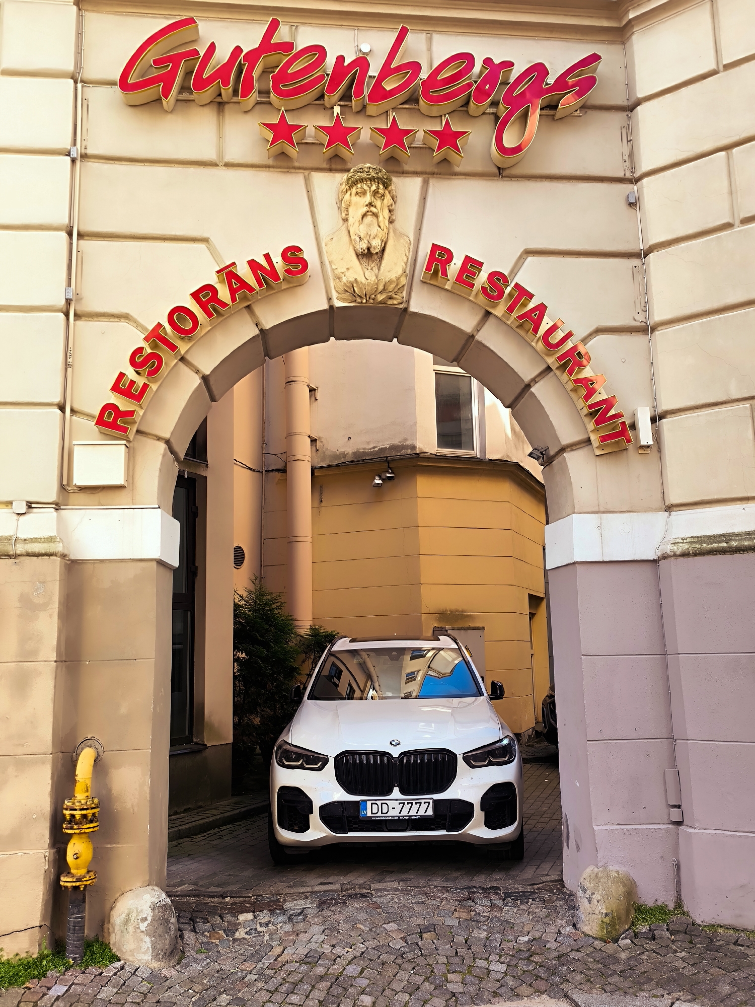 The image shows the entrance to the Gutenberg Hotel and Restaurant in Riga, Latvia. The hotel's sign, "Gutenbergs," is displayed in large red script at the top of the building. Below the sign, an archway frames the entrance, adorned with a bas-relief of a man's head and stars. The words "RESTORANS" and "RESTAURANT" are visible in red lettering on either side of the arch. A white BMW SUV is parked just inside the archway, facing the viewer. The ground is paved with cobblestones. A yellow gas pipe is visible on the left side of the entrance. The overall scene depicts a historic building entrance with a modern vehicle.