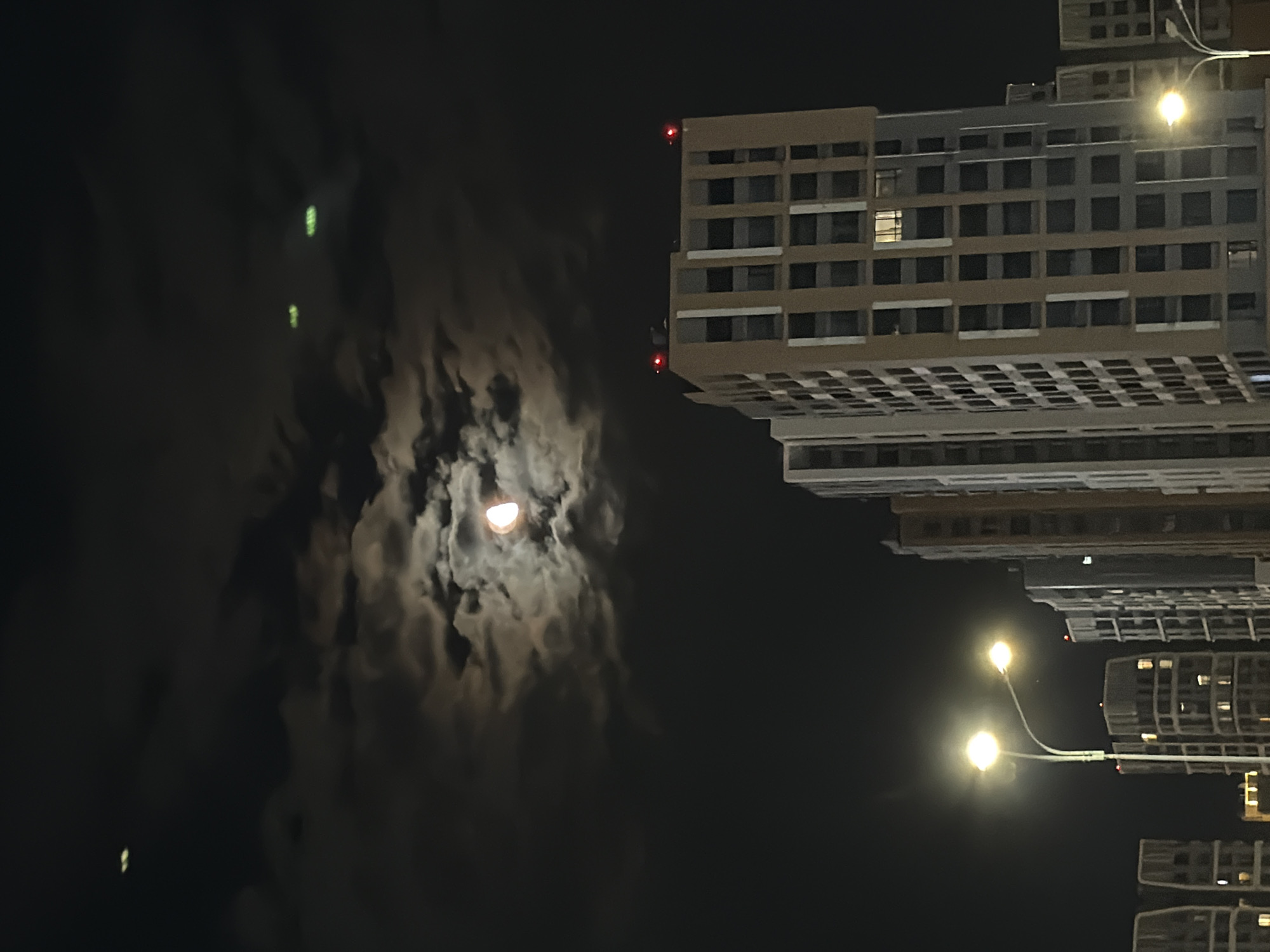 The image shows a nighttime scene in Astana, Kazakhstan, featuring a partially eclipsed moon partially obscured by clouds, and several modern residential buildings. The moon, with a crescent visible, is positioned in the upper center of the frame, illuminated by an unseen light source. Two distinct green lights are visible in the sky, one towards the upper left and another further to the right. The buildings are tall, multi-story structures with numerous windows, some of which are illuminated. Red lights, likely warning lights for aviation, are visible at the top of one of the buildings. A street lamp with two bright lights illuminates the lower left portion of the scene, casting a glow on nearby buildings and parts of the sky. The overall environment suggests an urban setting at night, with a mix of natural celestial phenomena and man-made structures.