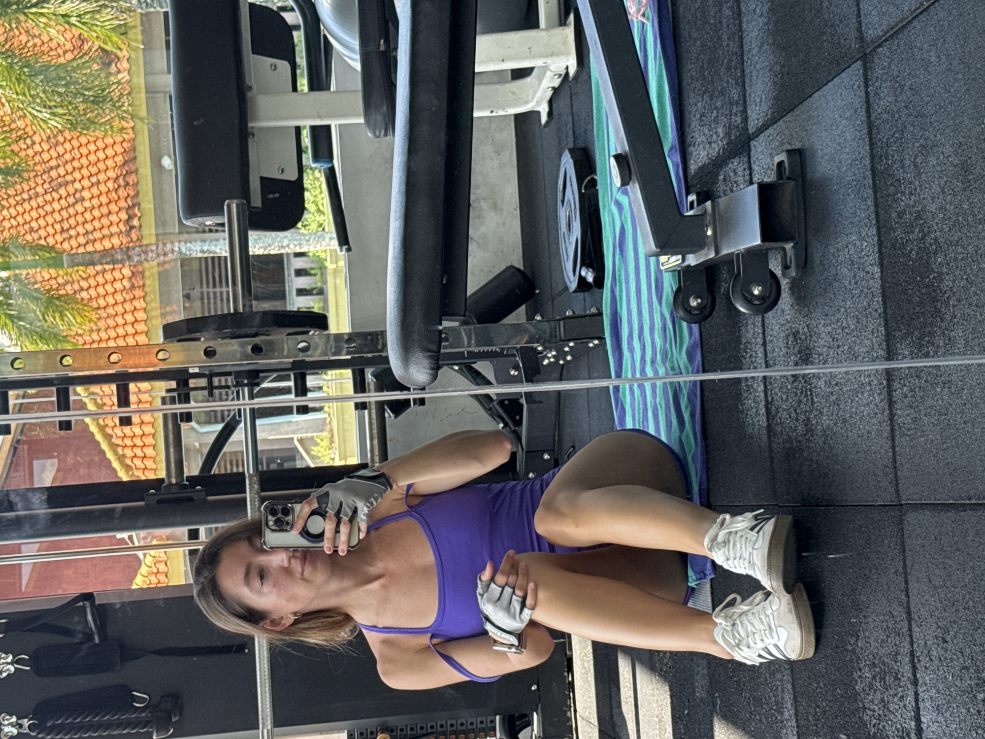 A woman is seated on a gym floor, taking a mirror selfie. She is wearing a purple tank top and shorts, white athletic shoes, and gray workout gloves. The woman has blonde hair and is holding a smartphone in her right hand. Behind her, a weightlifting bench, a barbell, and various gym equipment are visible. The gym appears to be outdoors, with palm trees and a building with orange tiled roofing visible through a large window. The setting is a gym located in Kuta Selatan, Bali, Indonesia.