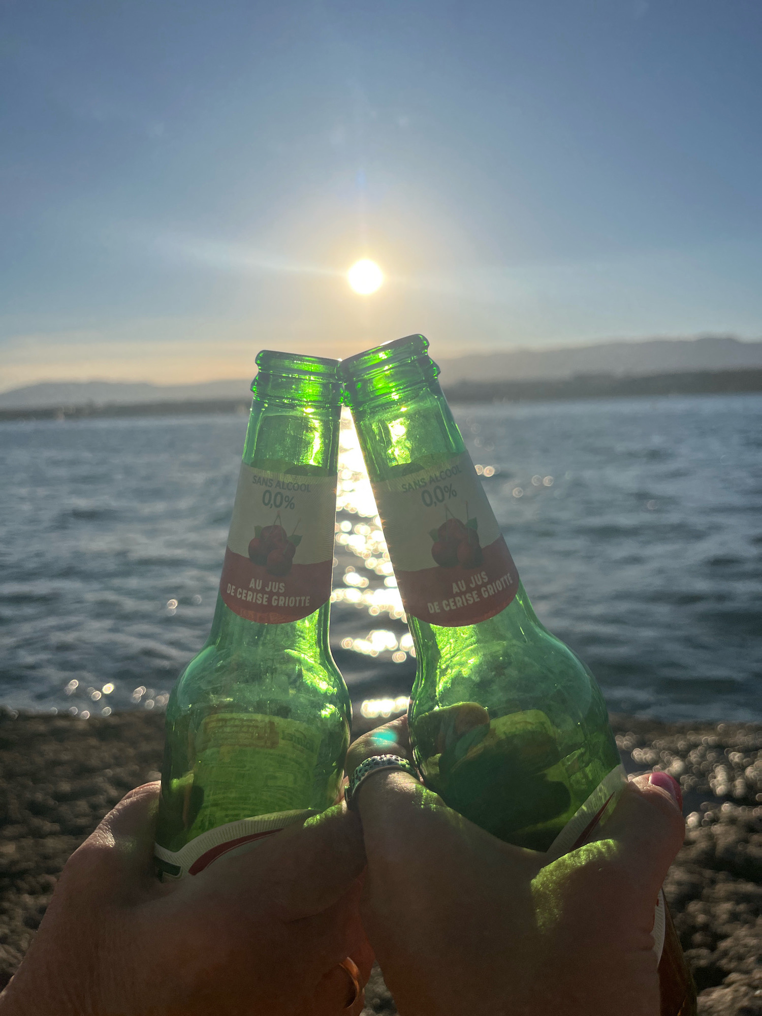 Two green glass bottles are held up in a toasting gesture against a backdrop of a sunlit body of water, likely Lake Geneva, with a faint mountainous horizon in the distance. The sun is positioned in the upper center of the frame, creating a bright glare on the water and illuminating the bottles. One bottle has a label that reads "SANS ALCOOL 0.0% AU JUS DE CERISE GRIOTTE," indicating it is a non-alcoholic drink made from sour cherry juice. The scene appears to be set in Cologny, Switzerland, given the prominent presence of Lake Geneva. The foreground shows parts of two hands holding the bottles, one hand wearing a ring and the other with painted fingernails. The ground beneath is rocky.