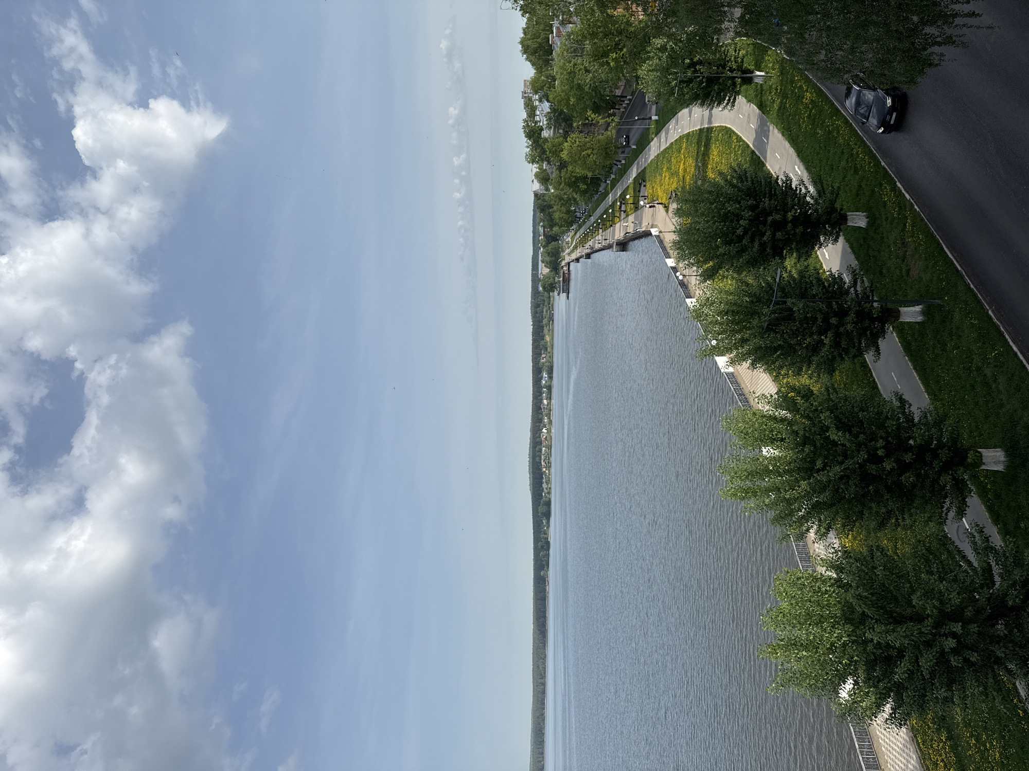 The image shows a body of water, likely a reservoir or wide river, adjacent to a paved embankment or promenade in Votkinsk, Russia. Along the waterfront, trees line the embankment, with some yellow flowers visible. A road runs parallel to the embankment, with a black car traveling on it. The sky is partly cloudy, with blue sky visible. The image is taken from a high vantage point.