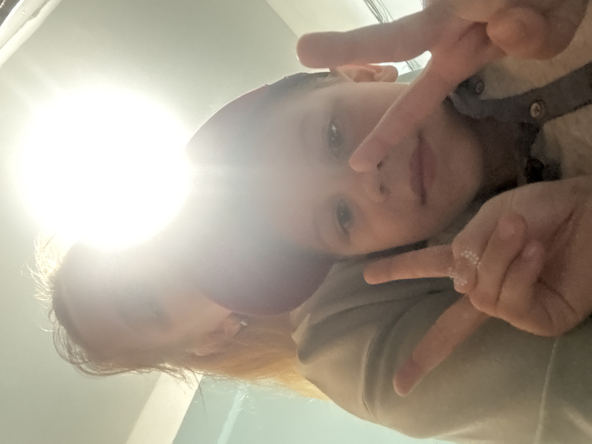 This is a close-up, low-angle selfie of two individuals, likely a mother and child, in what appears to be an indoor setting. The bright light source, possibly a window or lamp, is positioned in the upper left, causing significant overexposure and lens flare.
The foreground subject is a child, whose face is partially obscured by their own hand and the hand of another person. The child's eyes are visible, looking towards the camera. They are wearing a light-colored shirt or sweater with a dark buttoned placket. The other individual, presumably an adult, is positioned behind and to the side of the child, their head and shoulder visible. They have blonde hair and are wearing a red or pinkish head covering. Their hands are also visible, with one holding up two fingers in a peace sign near the child's head, and another hand resting on the child's shoulder. A ring with small stones is visible on one of the adult's fingers.
The individuals appear to be interacting closely, suggesting a moment of affection or playful posing for a photograph. The overall mood seems casual and intimate. The background is largely indistinct due to the lighting and shallow depth of field, but hints of indoor surfaces and possibly some clothing are discernible. No text is visible in the image. The lighting suggests it could be daytime, but the indoor setting makes it difficult to determine the exact time of day or weather conditions outside. The location context provided is Rusanovo, Russia.