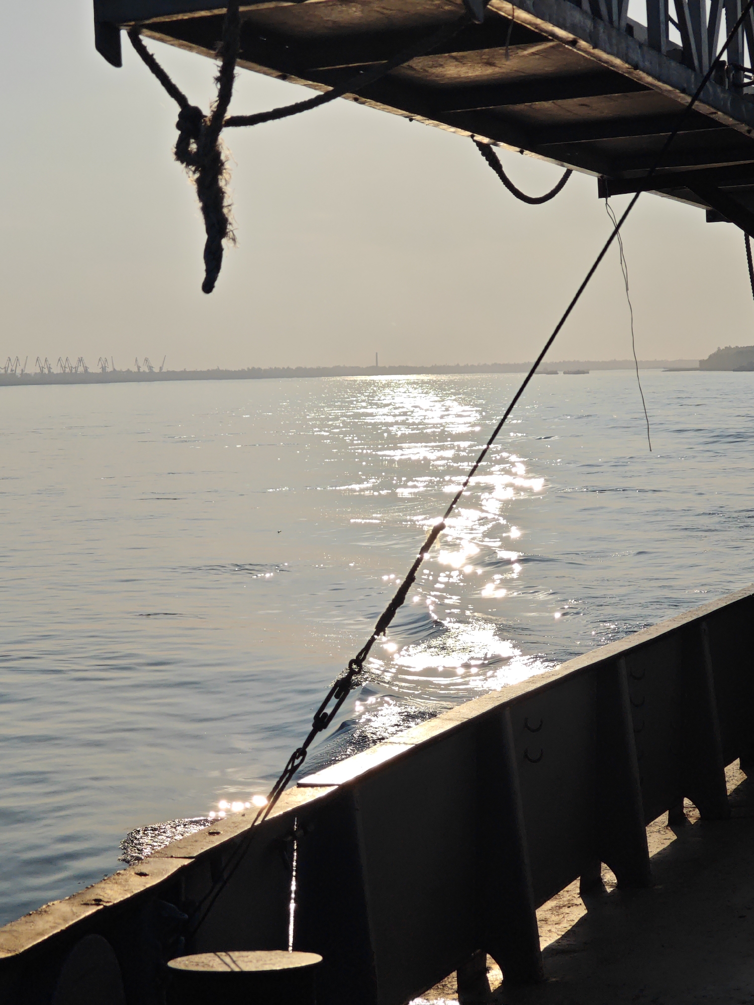 The image depicts a view from a vessel at sea, likely a ferry or cargo ship, looking towards the horizon. The sun is low in the sky, casting a shimmering reflection on the water. In the distance, a hazy shoreline is visible with industrial cranes and structures, suggesting a port or industrial area. A thick rope is hanging from a structure overhead on the left, and a taut rope with a chain is visible running diagonally across the scene from the foreground towards the right. The water appears calm with gentle ripples. The perspective is from a low angle, close to the water line, emphasizing the vastness of the water body and the open sky. The lighting suggests either early morning or late evening. The image does not contain any identifiable people. There is no indication of the specific location within the city of Lesosibirsk, Russia, as the scene depicts a maritime environment.
