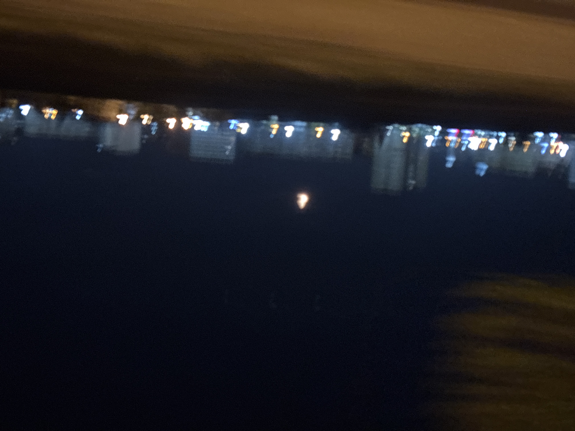The image shows a blurred view of city lights reflected in water at night. The lights likely belong to buildings, creating a shimmering effect. The location appears to be in Tbilisi, Georgia, evidenced by the distinctive urban layout and architectural characteristics. The foreground is dark and likely obscures the water's surface. The composition lacks specific details about individuals or interactions, emphasizing the abstract visual of the city lights.