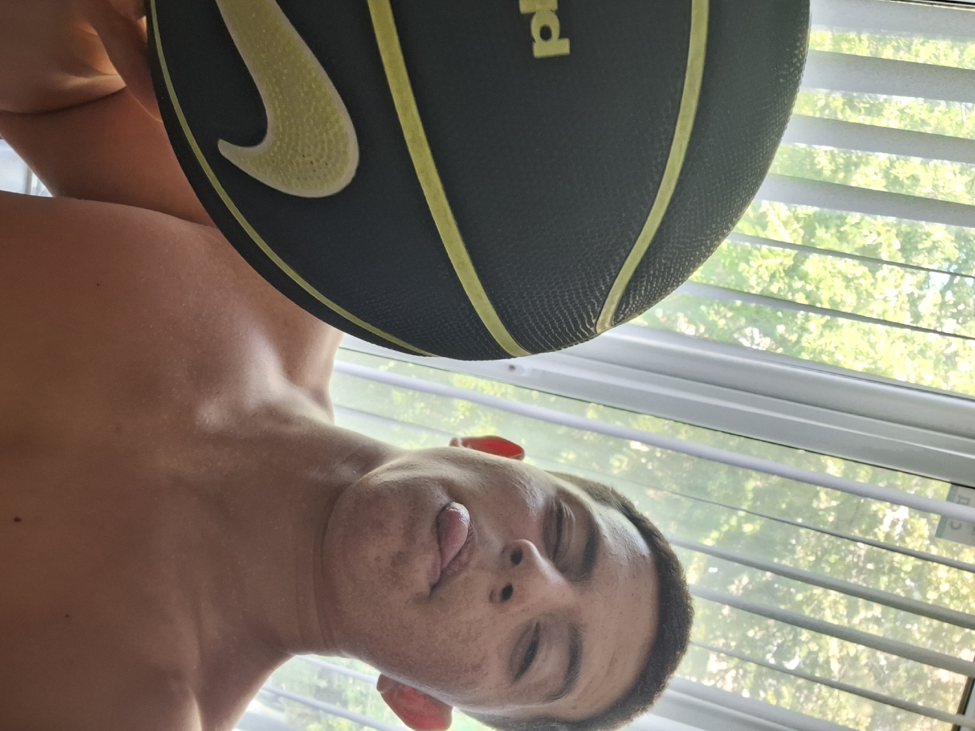 A young man with short, dark hair, shirtless, holds a black and yellow basketball to his left cheek. He is sticking his tongue out and winking. He has some blemishes on his face. The background appears to be a window with vertical blinds and trees visible through them, suggesting an indoor setting with natural light. The image does not provide any specific location information for Varna, Bulgaria.