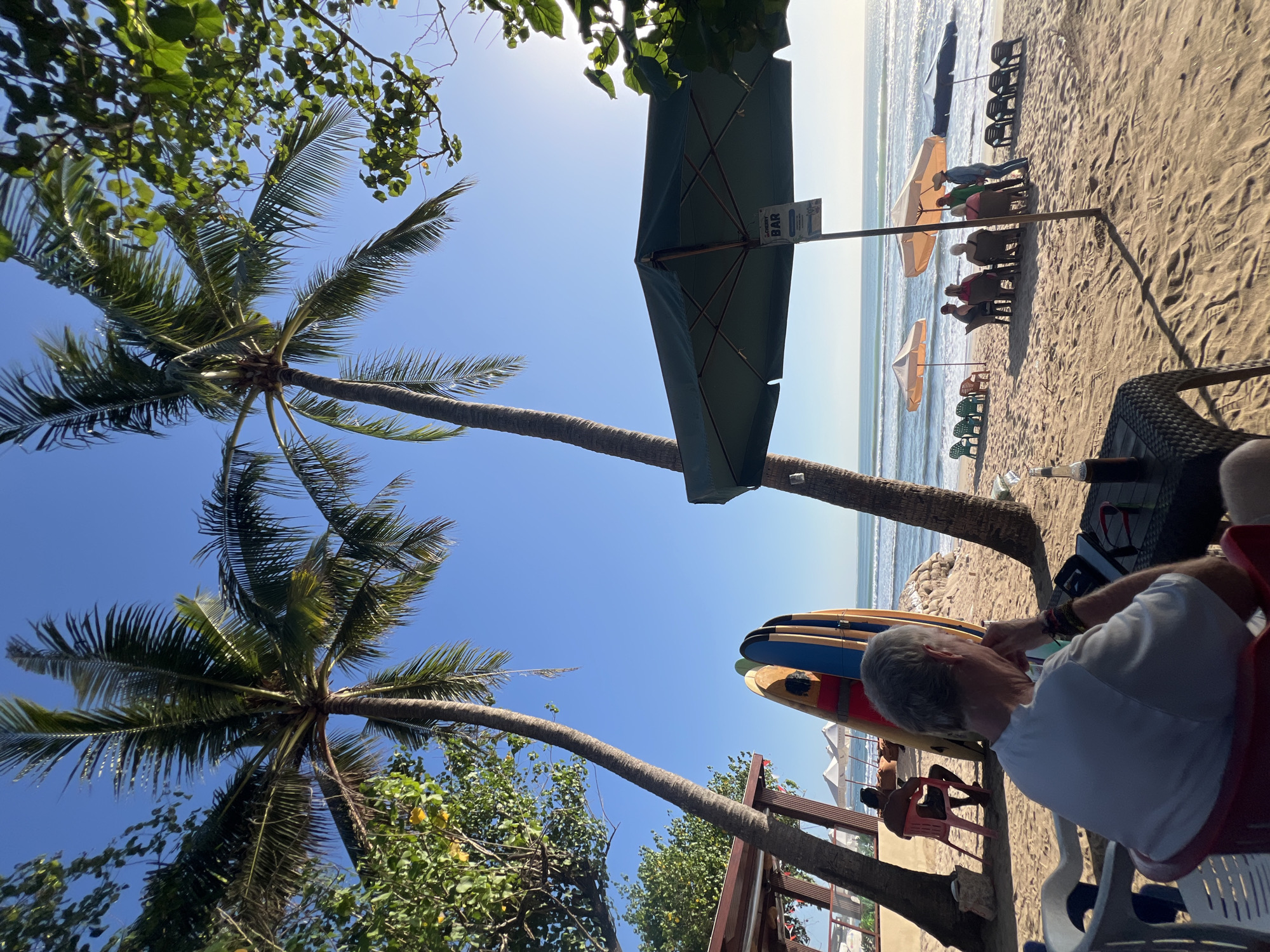 This is a photograph of a beach in Kuta, Indonesia, on a sunny day. In the foreground, an older man with grey hair is sitting in a red chair, facing away from the camera. He is wearing a white t-shirt and has his hand on his chin. On the table in front of him is a bottle of beer and a phone.
Behind him, a collection of surfboards are propped up. Further in the background, several people are seated at tables under umbrellas on the sandy beach, facing the ocean. The ocean has small waves, and the sky is clear blue. Palm trees are visible on the left and right sides of the image, with lush green foliage in the upper portion. A sign on a pole in the center reads "CHERRY BAR". The overall scene depicts a relaxed atmosphere at a beachside establishment.