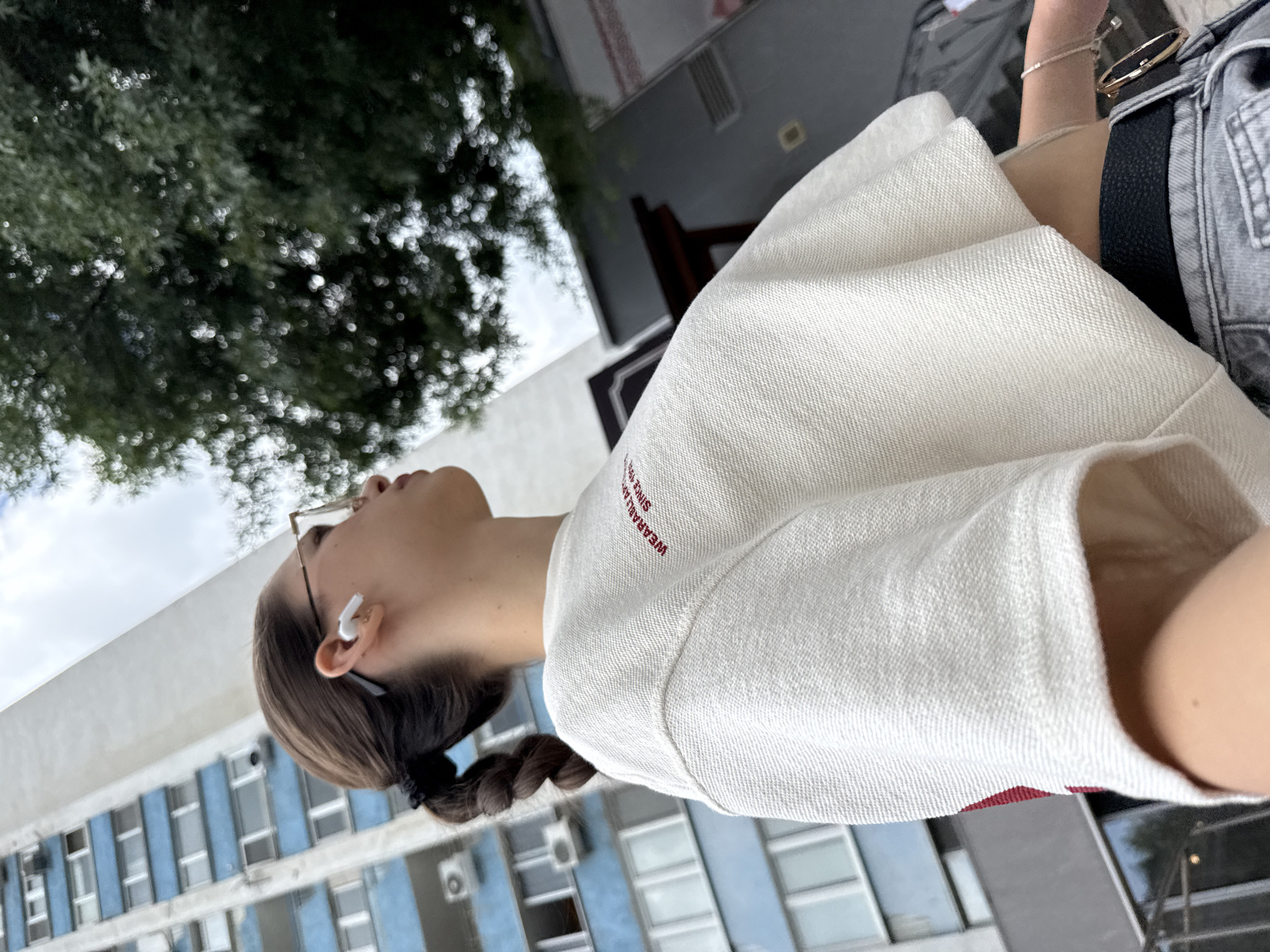 A young woman with brown hair pulled into a braid wears glasses and white earbuds. She is standing outdoors, facing away from the camera, looking upwards. She is wearing a white, cropped sweatshirt with red text that reads "WEARABLE ART SINCE 19". Her attire includes light-wash jeans and a black belt with a gold buckle. In the background, a tall, blue and white apartment building is visible, characteristic of Sofia, Bulgaria. A large, leafy green tree partially obscures the sky. The overall scene suggests an urban environment in Sofia.