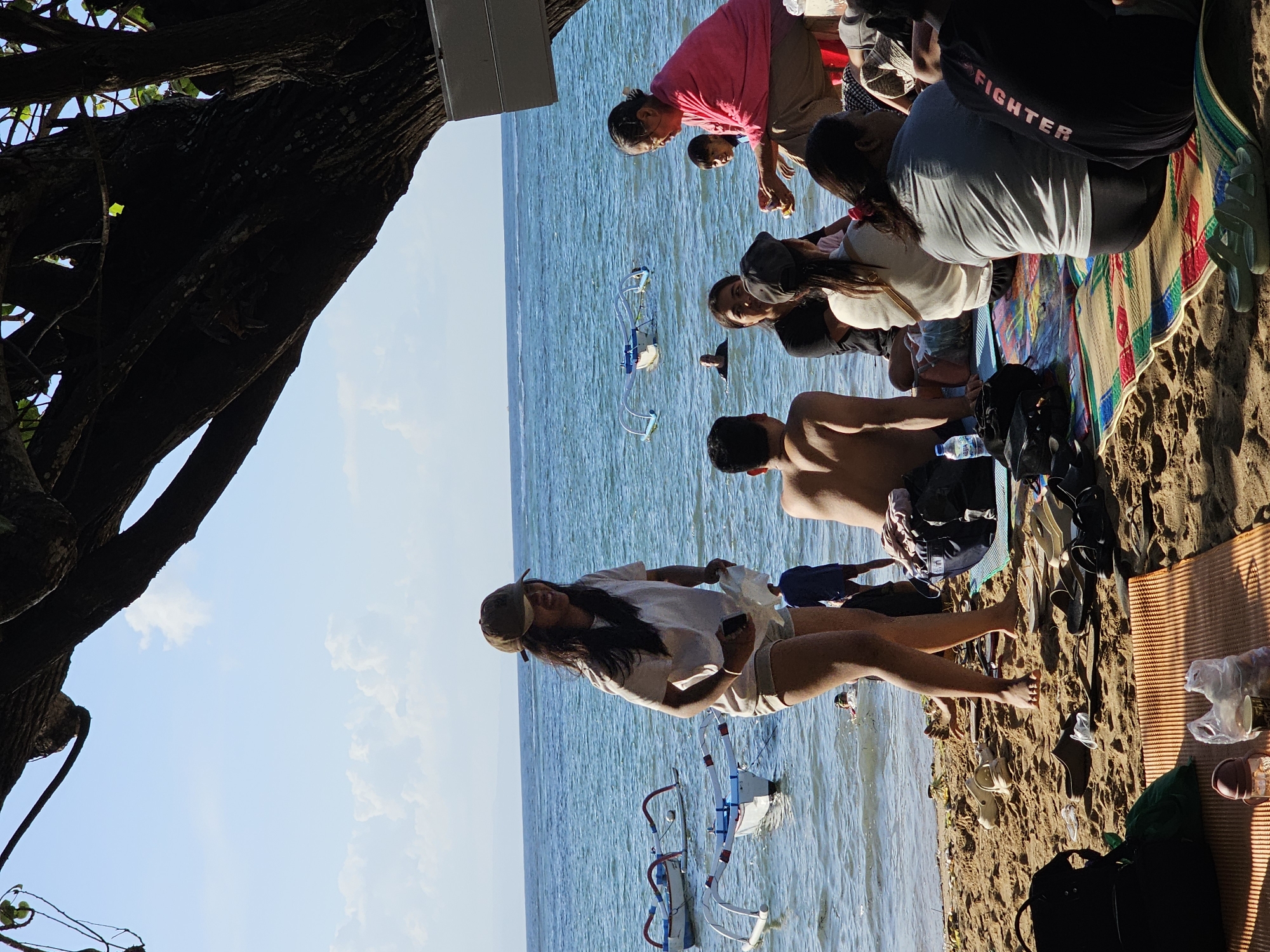The image depicts a beach scene in Denpasar, Indonesia, likely on a sunny day. In the foreground, several people are gathered on the sand, some sitting on mats. A woman in a light-colored t-shirt and shorts, wearing a cap, is standing and smiling near the water's edge. She is holding a phone and a white object. Several traditional Indonesian outrigger boats (Jukung) are visible in the shallow water. In the background, the ocean stretches to the horizon under a bright blue sky with scattered clouds. The presence of people, boats, and the general atmosphere suggest a leisure activity or a gathering at the beach. The sandy beach, the characteristic boats, and the tropical vegetation visible in the shadows of the large tree in the upper portion of the image all point to a location in Indonesia, specifically a coastal area like Denpasar.