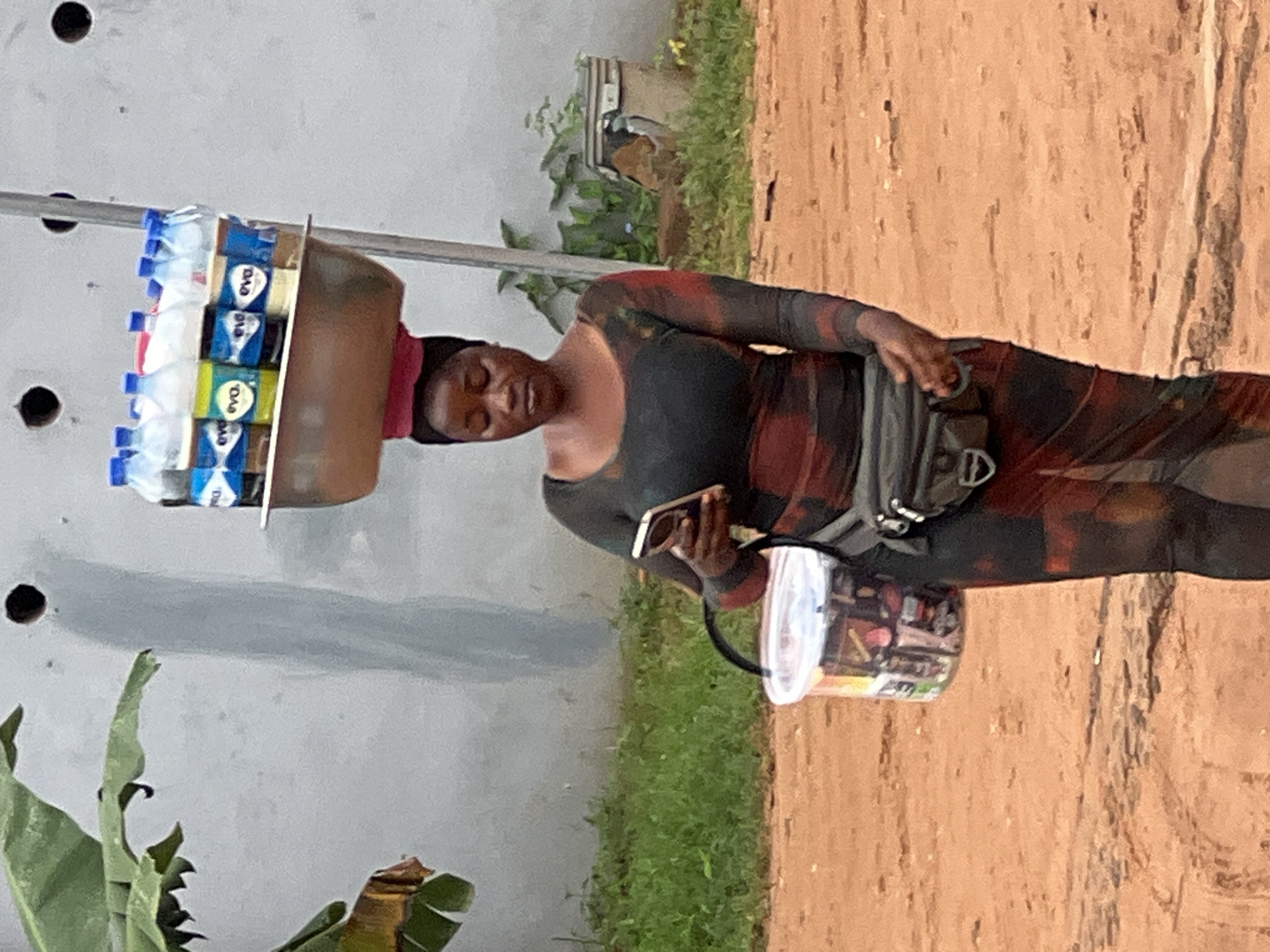 A Nigerian woman is pictured balancing a metal bowl filled with bottled beverages on her head. She is wearing a long-sleeved, form-fitting dress with a red and black pattern, and a black fanny pack around her waist. In her right hand, she holds a smartphone. A white bucket, containing various items, hangs from her left arm. The background consists of a grey wall, green vegetation, and a dirt ground. The scene appears to be on a street in Nkpor, Nigeria.