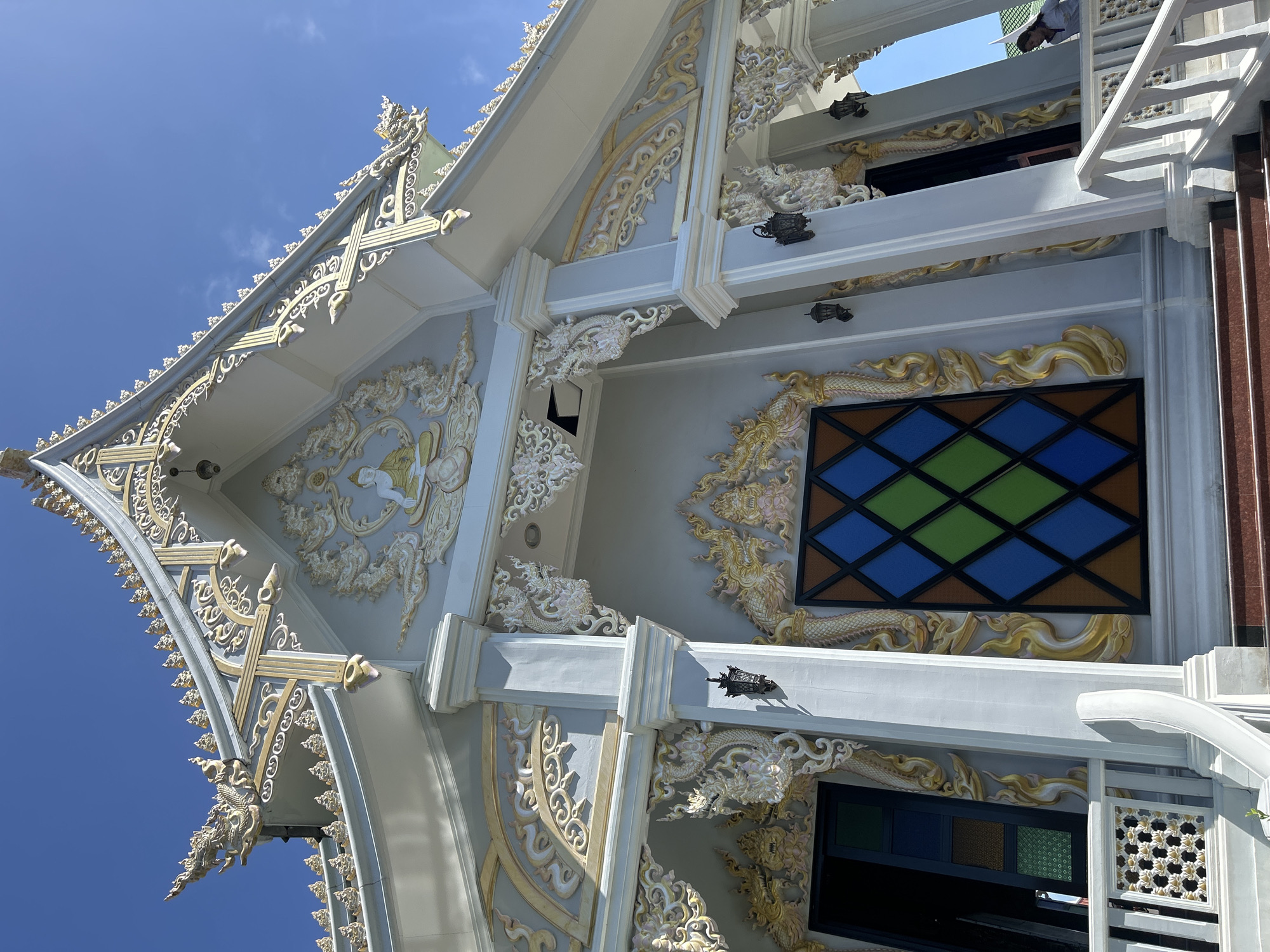 The image shows the exterior of a temple in Thai Ko Subdistrict, Thailand. The temple is light gray with ornate gold and white trim. The roof has intricate decorative elements, including what appear to be dragon or serpent figures and floral motifs. A Buddha image is depicted in relief above the entrance. The entrance features a stained-glass window with a diamond pattern in shades of blue, green, and brown. Two black lanterns are mounted on pillars flanking the entrance. White railings and stairs lead up to the temple. The sky is clear and blue. A person in a light-colored shirt is partially visible on the right side of the image, near the railings.