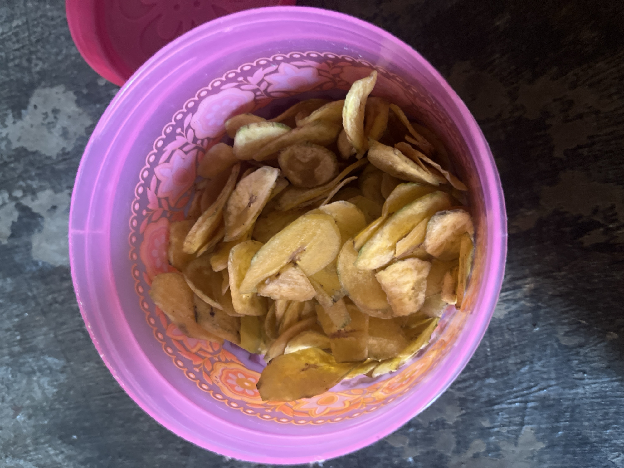 A close-up, overhead shot shows a pink plastic container filled with banana chips. The container has a decorative floral pattern around the rim. The banana chips are thin, golden brown, and appear to be sliced diagonally. The container is placed on a dark, textured surface that resembles wood or stone. A pink lid is visible to the right of the container, slightly ajar. The background is out of focus and shows a darker, mottled surface. The scene is likely indoors, possibly in a kitchen or dining area in Sidomulyo, Indonesia. There are no people or animals visible in the image.
