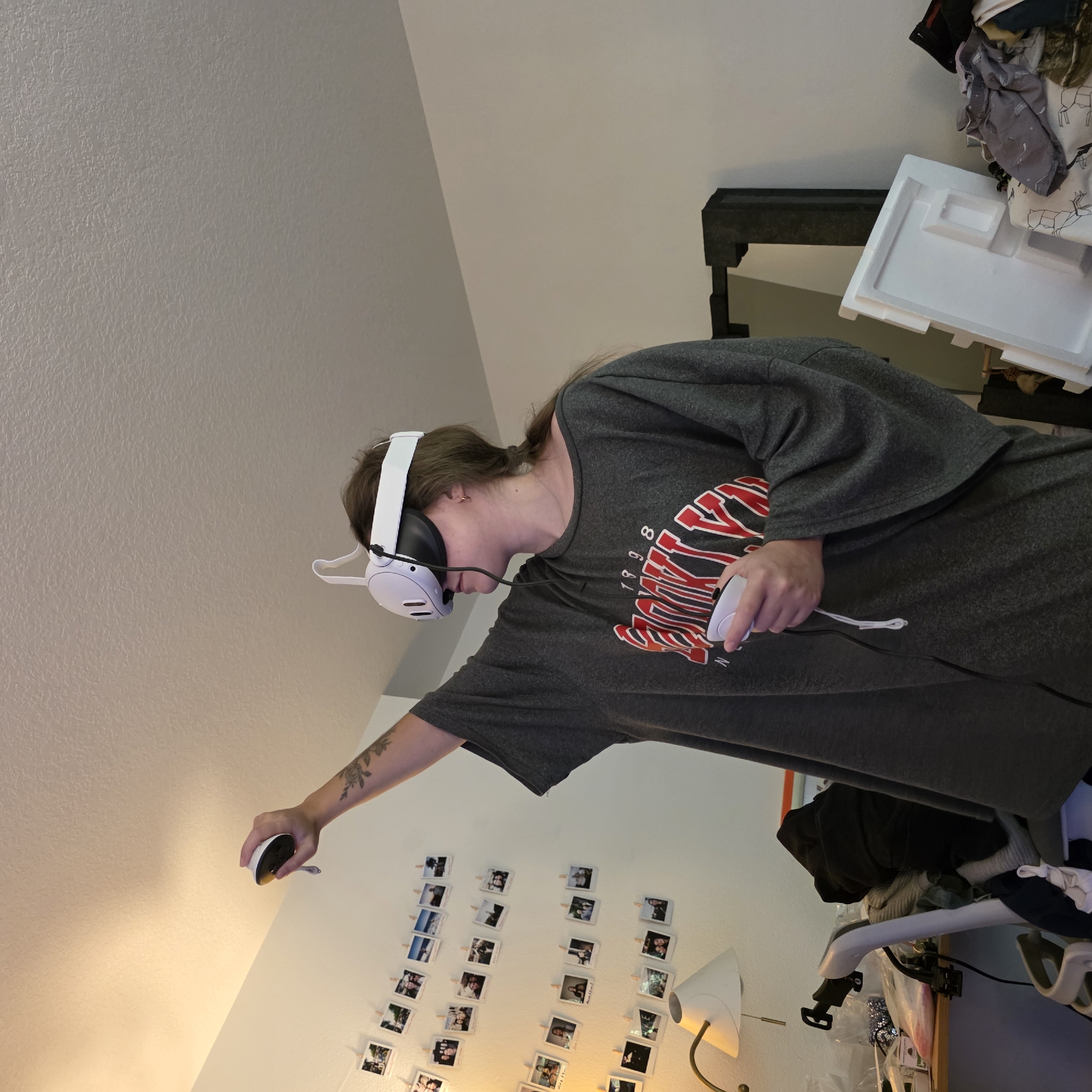 A person wearing a white VR headset and holding a VR controller is standing in a room, reaching their arm up. The person is wearing an oversized gray t-shirt with a red and white logo. The walls are white and decorated with numerous small photos clipped to strings. A lamp with a white shade is visible on a desk to the left. The room appears cluttered with clothing and other items.