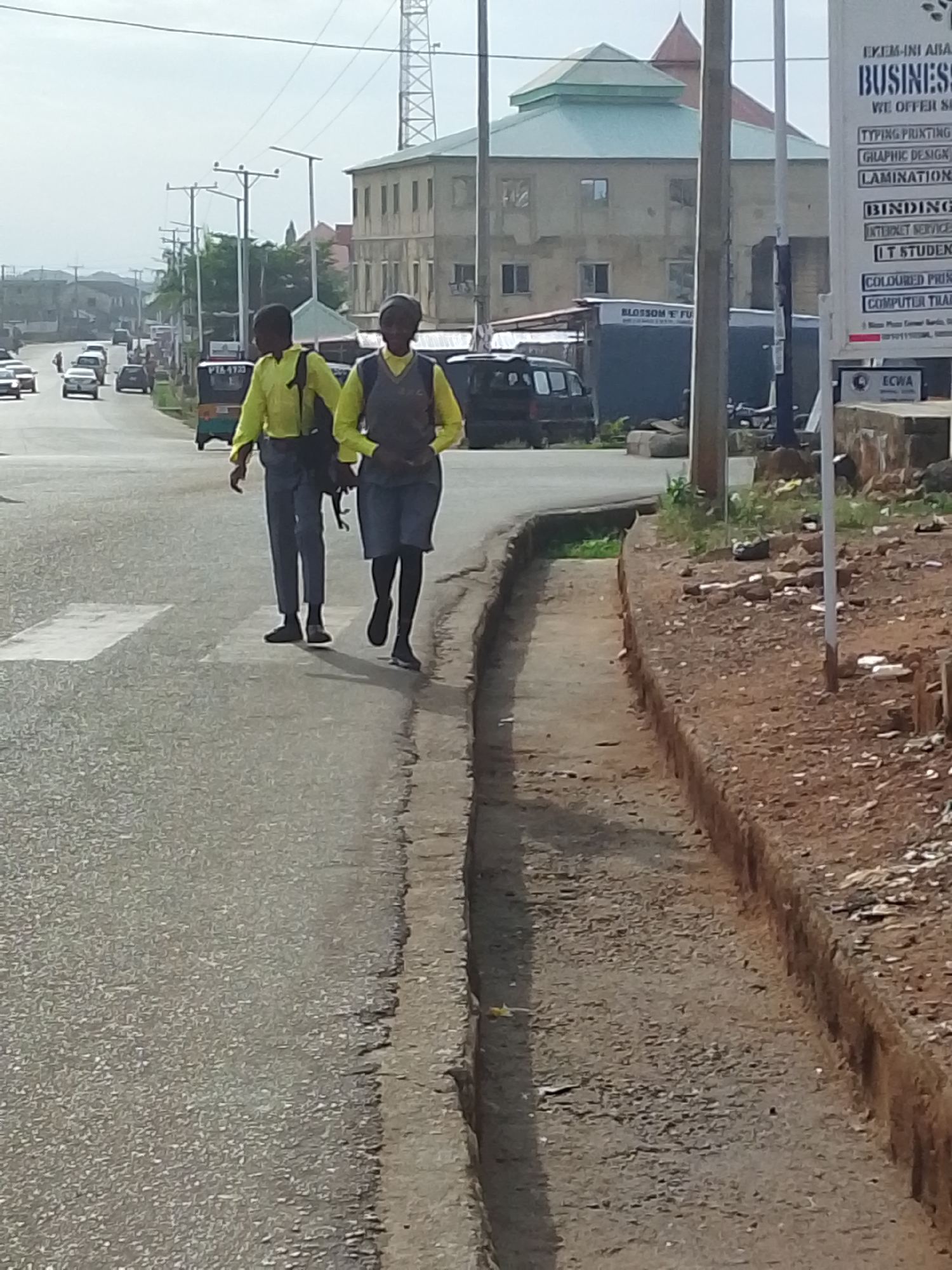 Two school-aged children in uniforms are walking along the left side of a paved road in Jos, Nigeria. They are walking in the same direction away from the camera, which is located on the opposite side of the road. The child closest to the camera has on a gray vest and dark-colored shorts with black leggings. The other child has on gray trousers. Both have on matching yellow long-sleeved shirts and dark colored shoes and book bags. A drainage ditch runs along the right side of the road, and the ground on the opposite side of the ditch is red and brown with scattered white trash. A sign stands on the right side of the road advertising business services. In the background, there are cars, a tricycle, and a building with a green rooftop. Overhead utility wires are present.