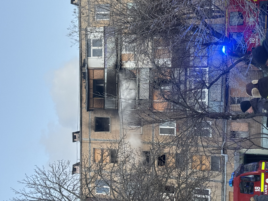 A multi-story brick apartment building exhibiting extensive fire damage is situated in the city of Unknown, Unknown. Active smoke emanates from multiple charred windows and a balcony on the mid-upper floors, specifically the fourth and fifth levels from the ground. Extensive charring is visible around affected window frames and across the building's facade. Bare deciduous trees partially obscure the lower and mid-sections of the building in the foreground. Two red fire trucks are partially visible at the base of the building; one on the left displays the number "18" and the text "ХАРКІВ" (Kharkiv), while another on the right features flashing blue emergency lights. Several individuals, dressed in winter attire, stand near the emergency vehicles. A visible stream of water or foam is being directed towards the building, indicating active fire suppression efforts. The sky is partly cloudy and blue, consistent with daytime conditions.