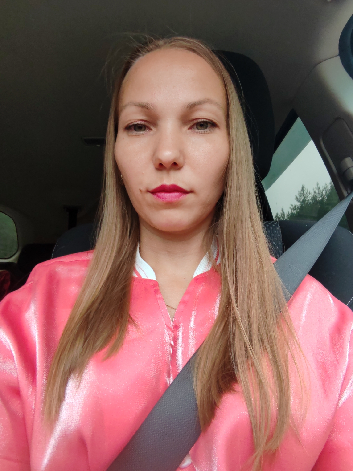 A selfie is taken from the chest up of a Caucasian woman with blonde hair, wearing a bright pink jacket and a gray seatbelt across her chest. She has red lipstick on and her eyes are open, looking directly at the camera. She appears to be in the driver's seat of a car, with the car's interior visible in the background, including a headrest, a window with trees visible outside, and the roof of the car. The lighting suggests it is daytime. There is no visible indication of the city of Yb, Russia.