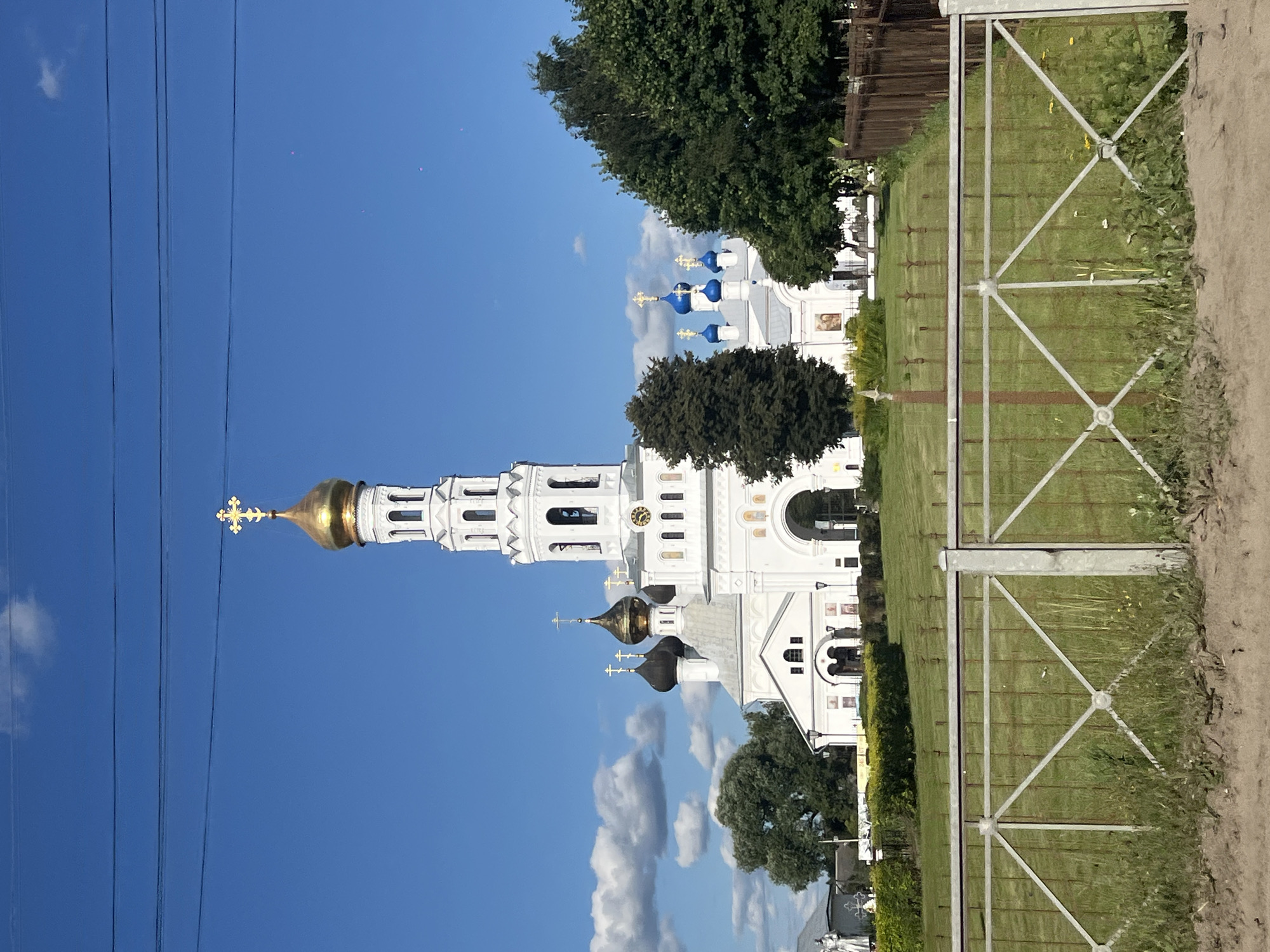 The image shows a large white Orthodox church with a tall bell tower topped by a golden dome and cross. The church has multiple domes, some black and some blue, also topped with crosses. A clock is visible on the bell tower. The church is surrounded by a well-maintained green lawn and trees. A white fence with a decorative metal pattern runs across the foreground of the image. In the background, there are more trees and a clear blue sky with some white clouds. The church is identified as being in the city of Zavidovo, Russia. There are no people or animals visible in the image, and no actions are taking place. The environment is sunny and appears to be summer or early autumn.