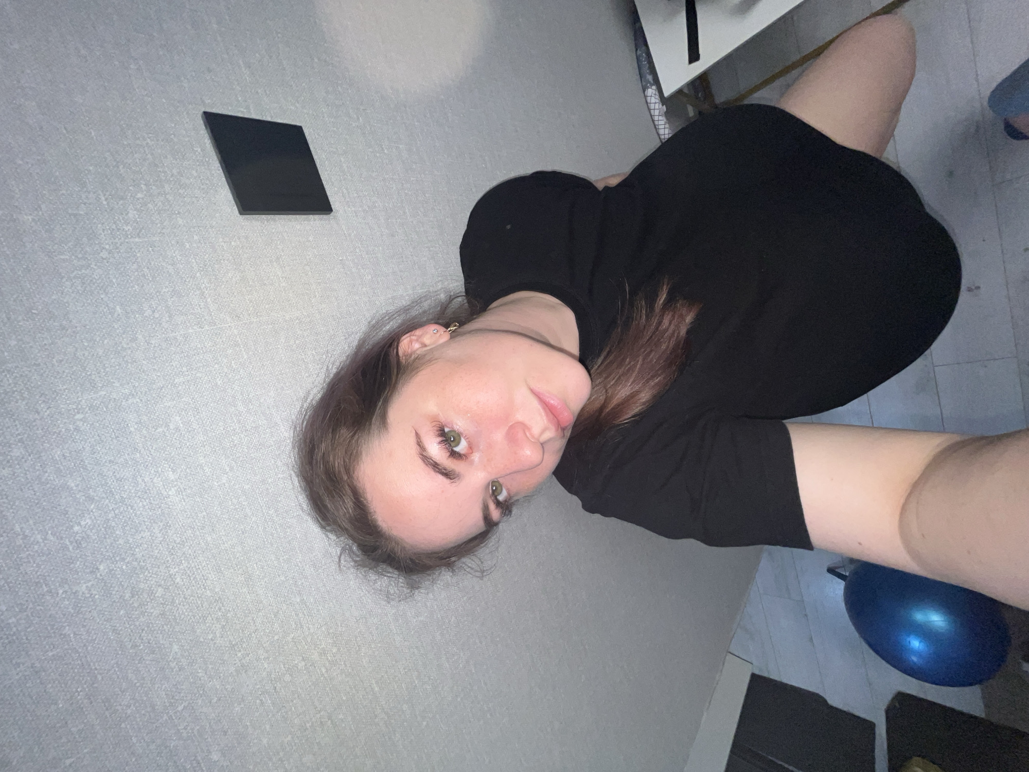 A young woman is lying down on a floor near a wall. She is wearing a black dress. A black rectangular object is on the wall. A blue exercise ball is visible near her legs. A white desk is partially visible. The location is unknown and there is no information to indicate the city of Chaikovsky, Russia.