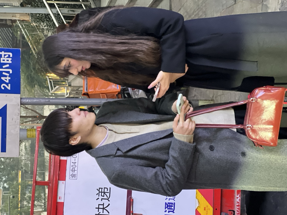 Two individuals are depicted in an outdoor urban setting in Yuzhong District, China. The person on the left, appearing male, has dark medium-length hair with bangs and wears a small earring in the left ear. He is dressed in a grey double-breasted coat over a light beige collared knit sweater, with a subtle light purple trim visible at the neckline. He holds a light green smartphone in his right hand and carries a glossy reddish-orange shoulder bag by its strap in his left hand. He is looking towards the person on the right.
The person on the right, appearing female, has long, dark, wavy hair with bangs. She wears a long, dark, full-length coat. She holds a white smartphone with a pink case in her left hand, and her fingernails are painted pink. She is looking towards the person on the left. Both individuals are in direct eye contact and are in close proximity.
In the background, to the left of the male-presenting individual, a red and white delivery vehicle or cart is partially visible, displaying Chinese characters "快递" (express delivery) and "渝中04-00" (Yuzhong 04-00), along with a "3M" logo. A vertical metal pole with a blue rectangular street sign is positioned between the two individuals. The sign features a white arrow pointing left and the Chinese characters "24小时" (24 hours) in white. To the right of the female-presenting individual, a set of concrete steps with a silver metal railing is visible, along with some green foliage above. Partially seen orange delivery bags are also present in the background. The scene is illuminated by diffused natural light.