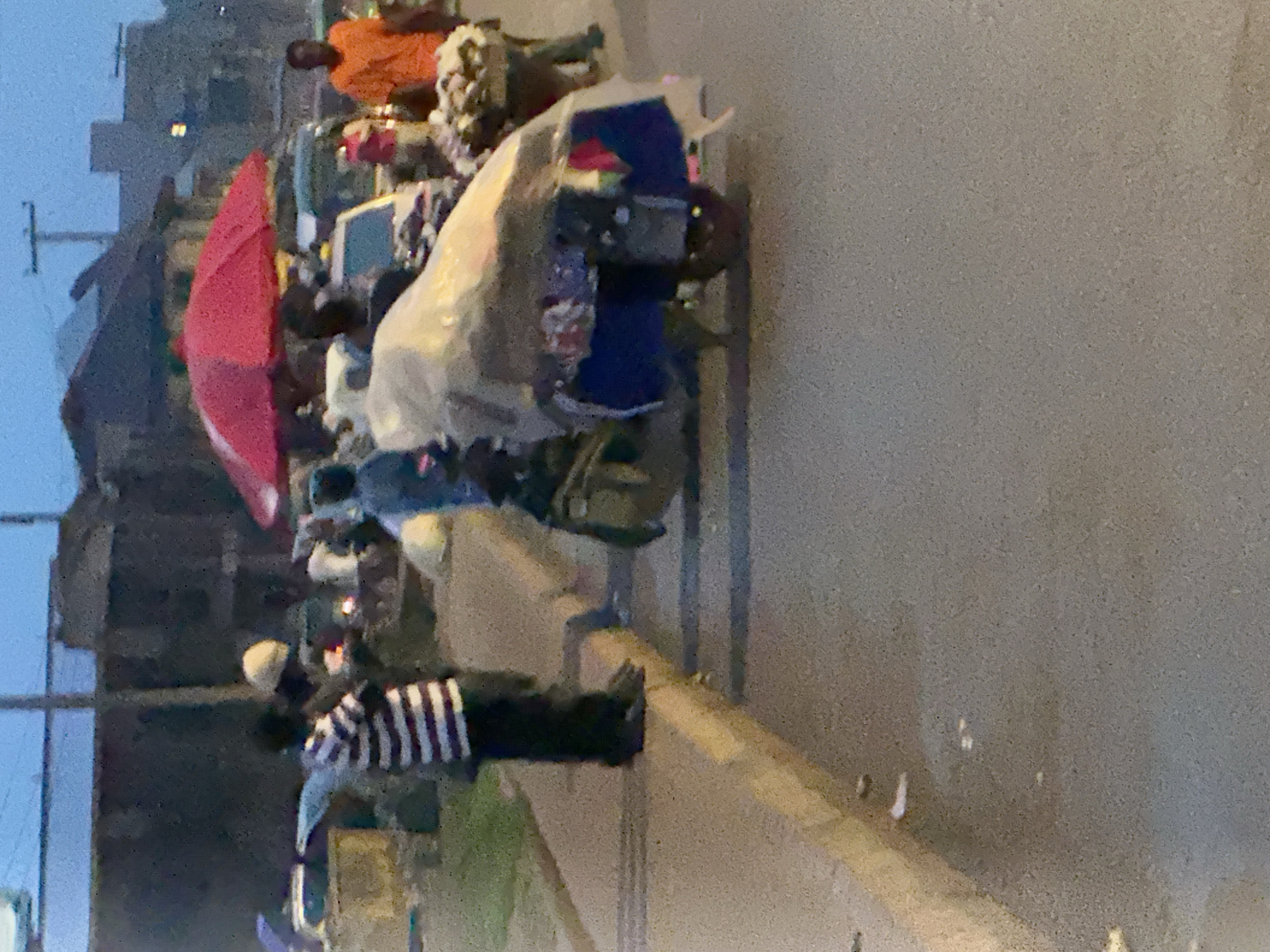 In Bukuru, Nigeria, a street vendor stands beside a motorcycle laden with goods covered by a tarp. The vendor wears a black and white striped shirt and dark pants. The motorcycle is adorned with a blue cloth and what appears to be an assortment of items, including fabric. A red umbrella is open over the motorcycle. In the background, other pedestrians and vehicles are visible, suggesting a busy urban environment. The sky is a dim twilight blue.