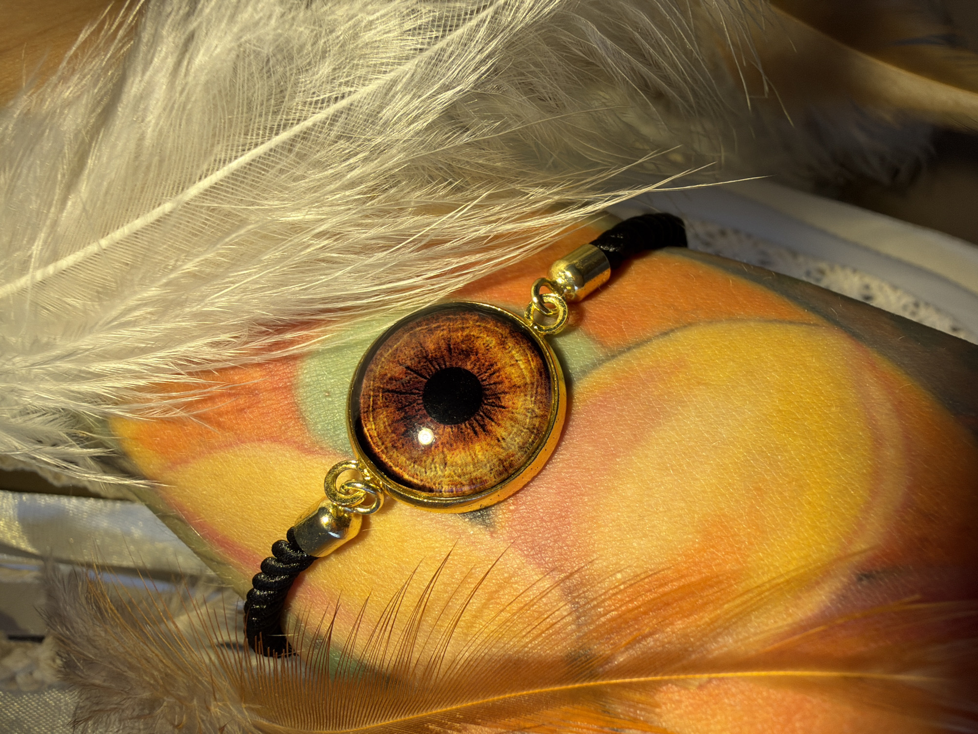 The image shows a close-up of a bracelet with a round, golden-rimmed pendant featuring a detailed iris design, likely from an eye. The bracelet itself is made of a black cord. It is placed against a textured surface with vibrant orange and yellow hues, possibly skin with a tattoo or colored fabric. Several feathers are arranged around the bracelet: two brown feathers on the left, with their delicate barbs visible, and a cluster of white feathers on the right, also showing intricate detail. The lighting creates strong highlights and shadows, emphasizing the textures of the feathers and the pendant. The background is blurred, suggesting a shallow depth of field. There is no discernible location in Nha Trang Ward, Vietnam, evident in this image.