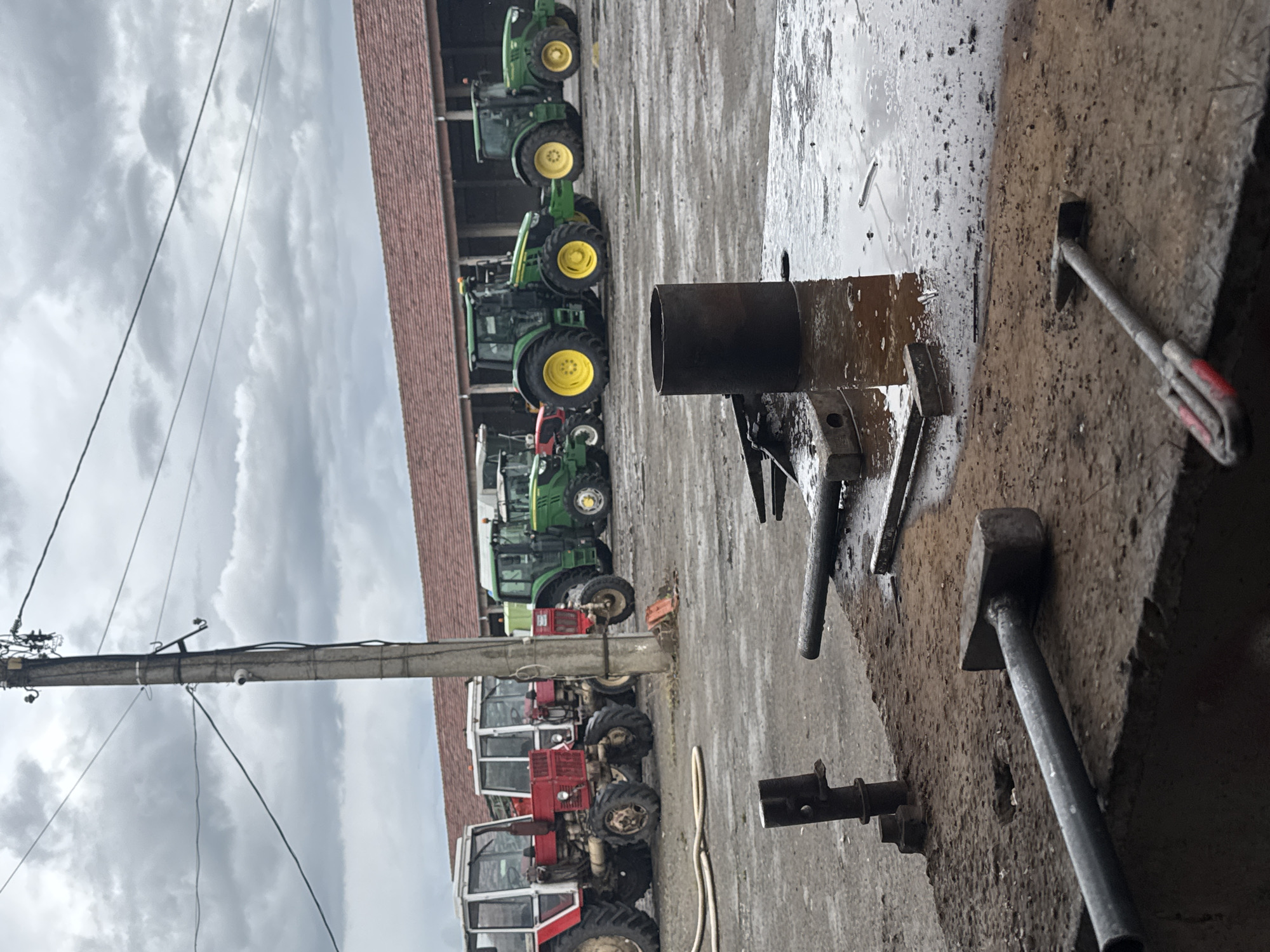 The image depicts an outdoor scene, possibly a farmyard or agricultural repair area, located in Sarnevo, Bulgaria. A row of tractors is parked along a building with a red tile roof. The tractors vary in model and color, with green John Deere tractors and red MTZ tractors visible. A utility pole is positioned near the row of tractors. In the foreground, a cluttered workbench area is present with tools, including a sledgehammer and other metal implements, scattered around. The area appears to be wet or recently rained on. The sky overhead is overcast with grey clouds. No people are visible. The focus is on the tractors and the repair area, suggesting a rural agricultural setting.