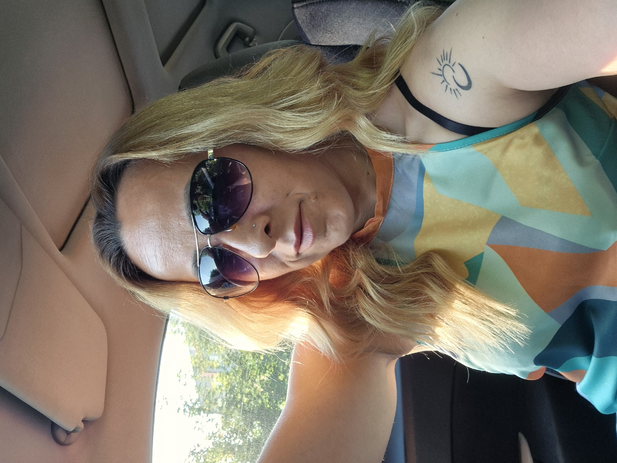 A woman with blonde hair and sunglasses is taking a selfie in a car. She is wearing a colorful, geometric-patterned top and has a tattoo of a sun and crescent moon on her left shoulder. The car's interior is visible, including the sun visor and ceiling. Sunlight streams in from the left, illuminating her face and hair. Trees and foliage are visible through the car window.