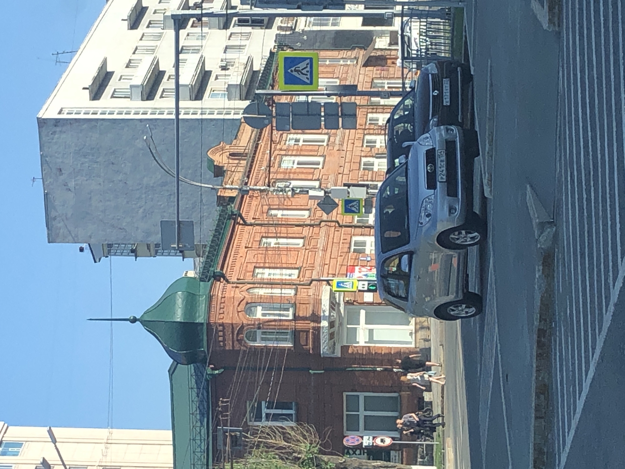 The image depicts a street scene in a city. In the foreground, a silver SUV is stopped at a crosswalk, with a dark-colored sedan behind it. The cars are on a paved road with white lines indicating a pedestrian crossing.
In the midground, a red brick building with a distinctive green domed roof is prominent. The building has multiple windows and architectural details. A pedestrian crossing sign with a walking person symbol is visible on a pole in front of the building. Several people are walking on the sidewalk in front of the building, some appearing to be conversing.
In the background, modern multi-story buildings rise behind the brick structure, some with balconies and numerous windows. The sky is clear and blue, suggesting it is daytime. The overall environment appears to be an urban setting with a mix of older and newer architecture.