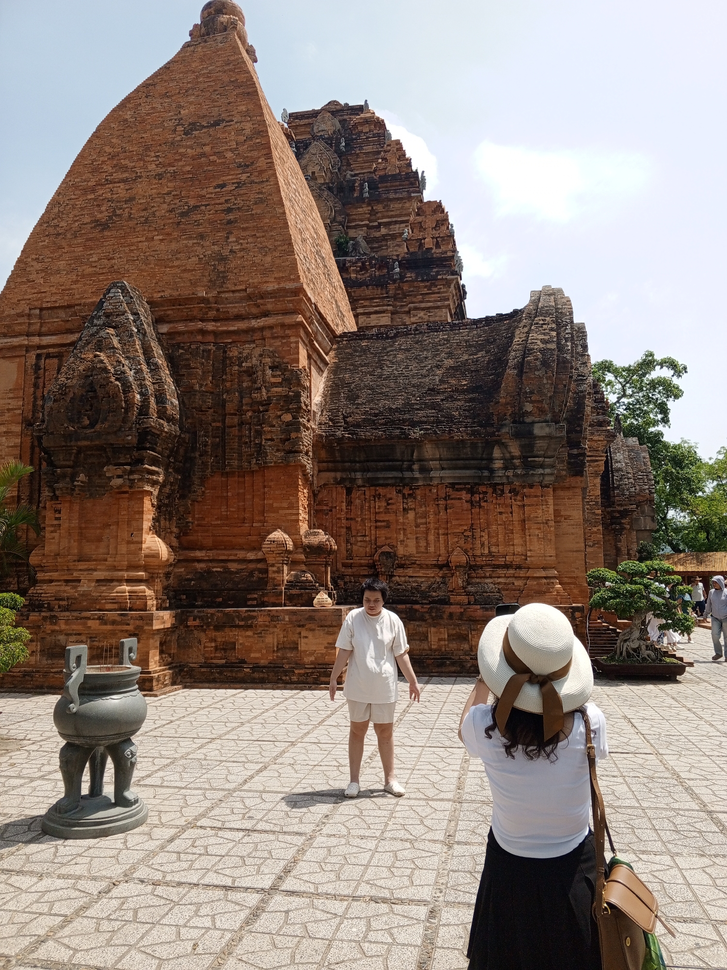 The image depicts the Po Nagar Cham Towers in the city of Nha Trang, Vietnam. In the foreground, a woman wearing a white shirt and a wide-brimmed hat with a brown ribbon is taking a picture of the towers. She has dark, curly hair and is wearing a dark skirt and a brown shoulder bag. To her left, another woman in a white t-shirt and white shorts is standing and looking towards the towers. In the mid-ground, the large, reddish-brown brick Cham towers are prominent, showcasing their intricate architecture. A large, dark, ornamental urn with four legs sits on the tiled ground to the left of the frame, filled with incense sticks. The ground is paved with hexagonal stone tiles. In the background, trees and other structures of the complex are visible, along with a few other people walking around. The sky is bright and overcast.